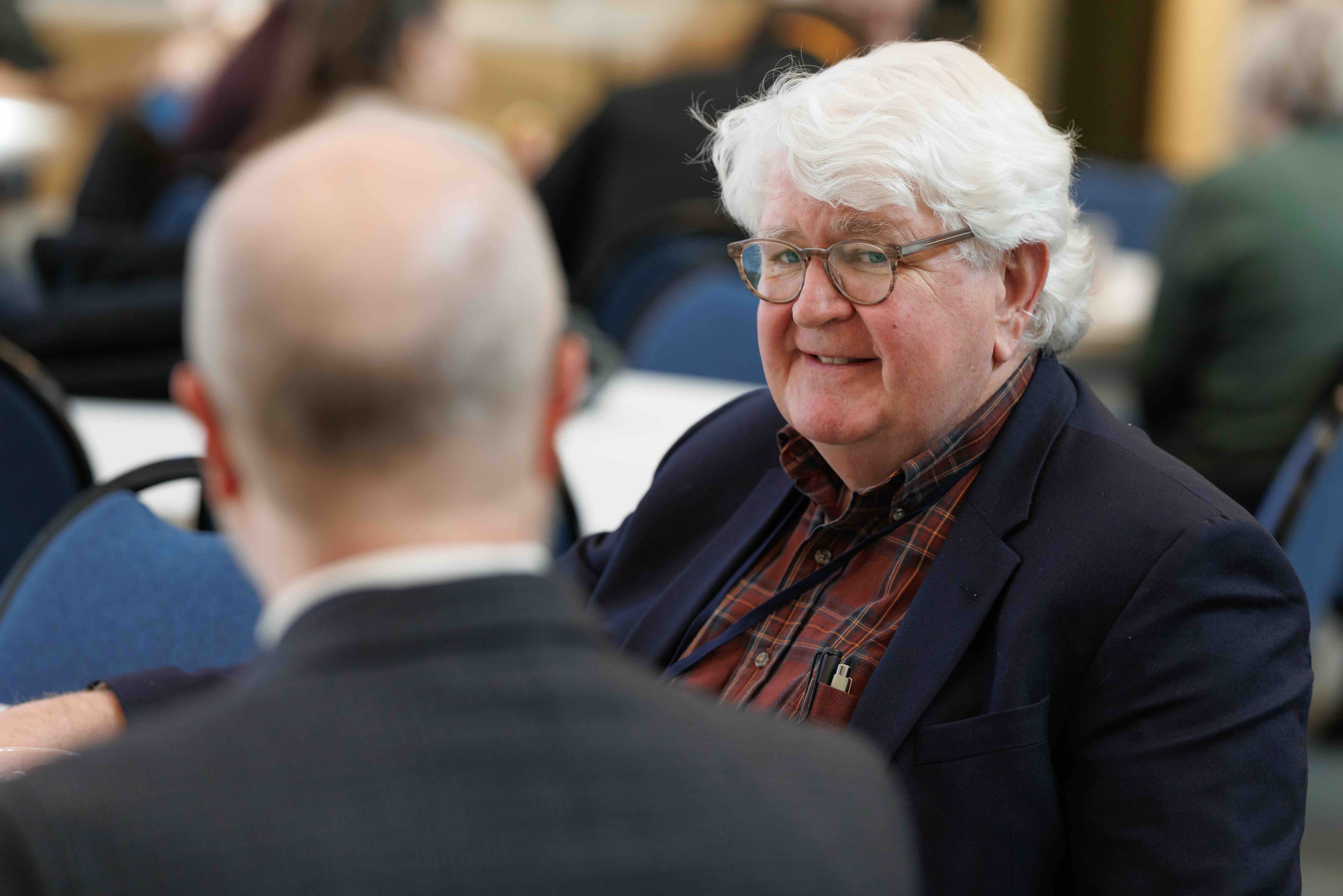 Two men are seated at a table conversing. The man facing the camera has white hair, glasses, and wears a dark blazer, appearing engaged and positive.