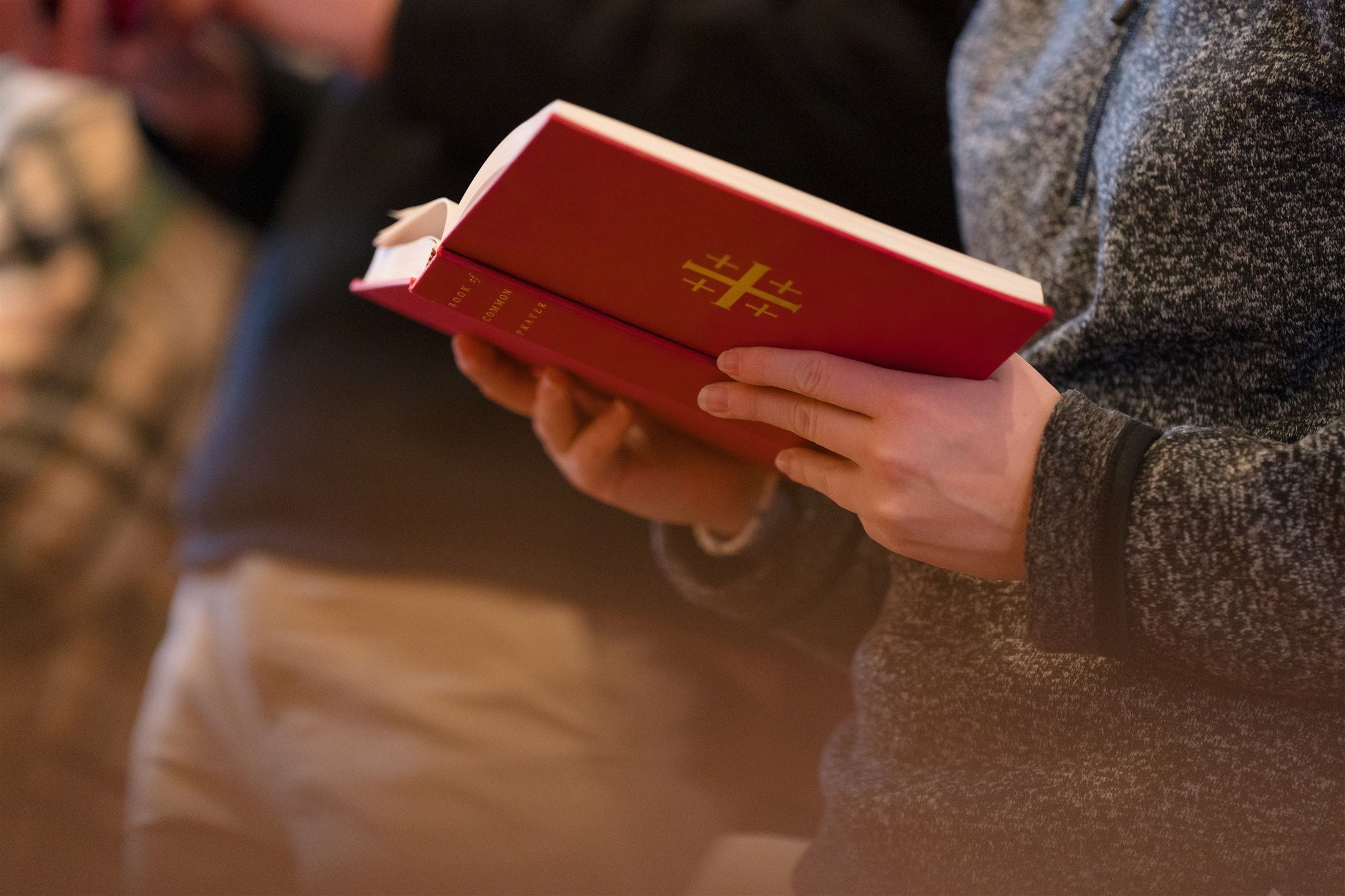 A person in a gray sweater holds an open red hymn book with a gold cross symbol during a church service, creating a peaceful, reflective mood.