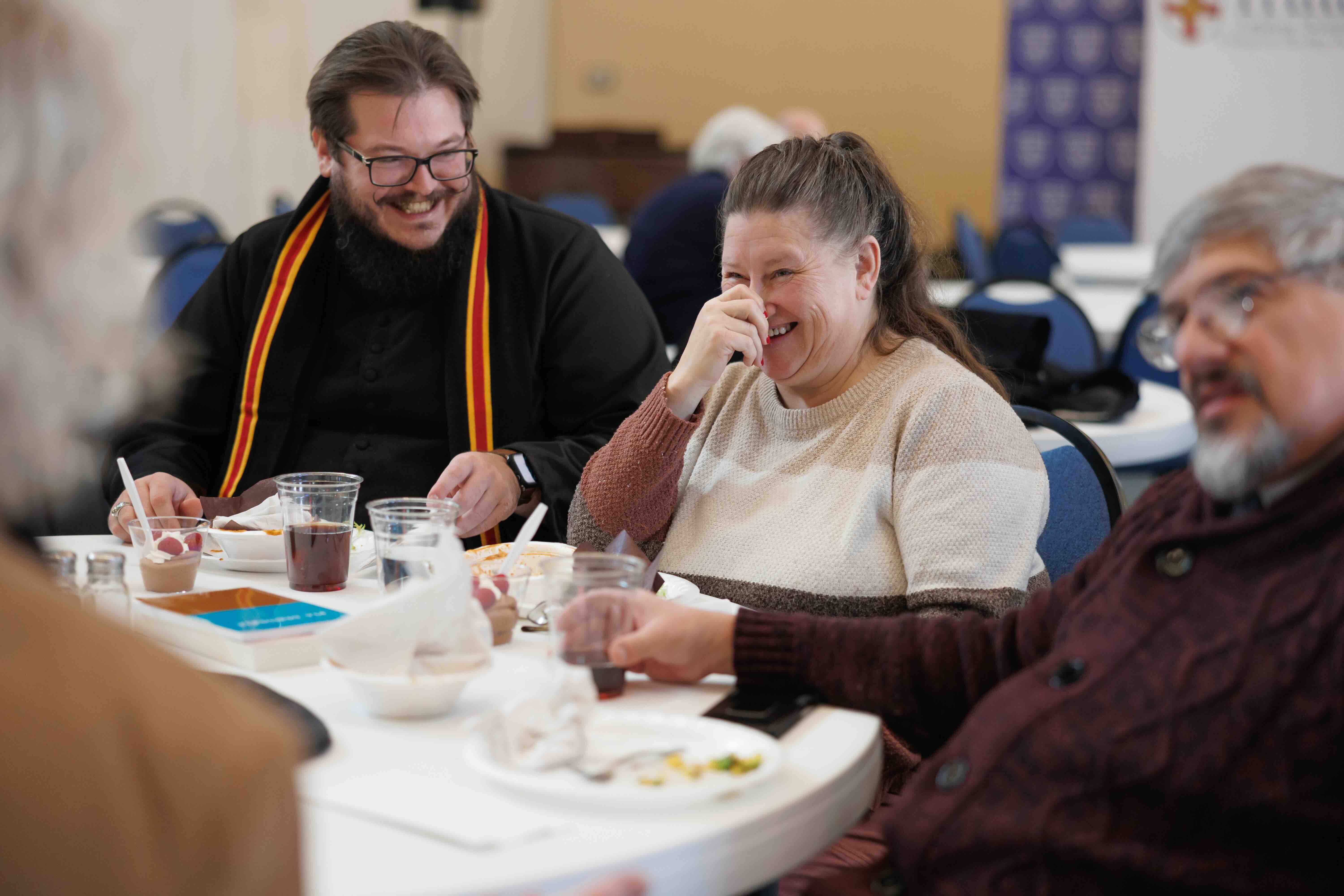 Three adults sitting around a table sharing a meal and laughing together in a casual setting.