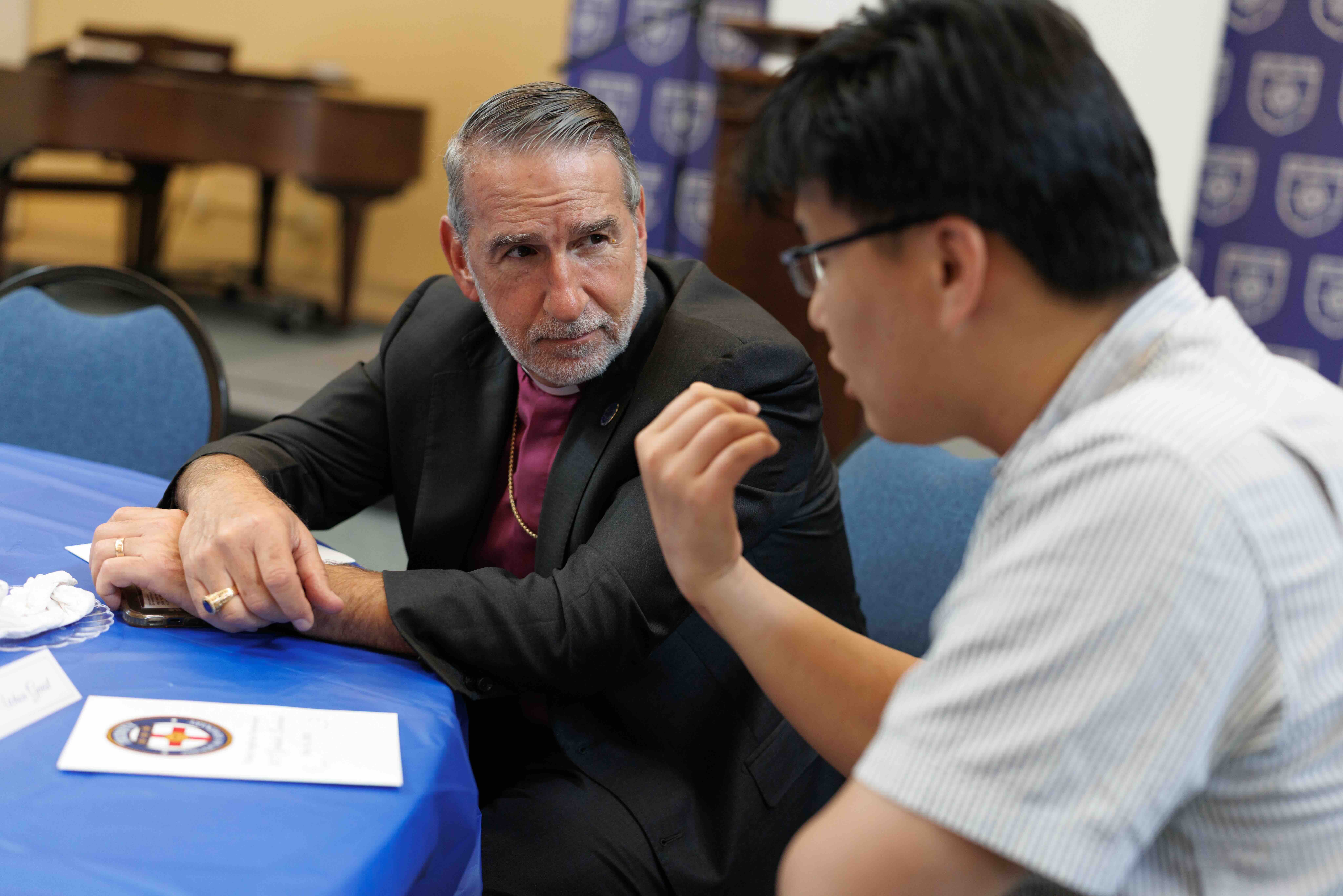 A man in a clerical collar listens intently to another man in glasses during a conversation at a blue-tableclothed table, conveying focus and engagement.