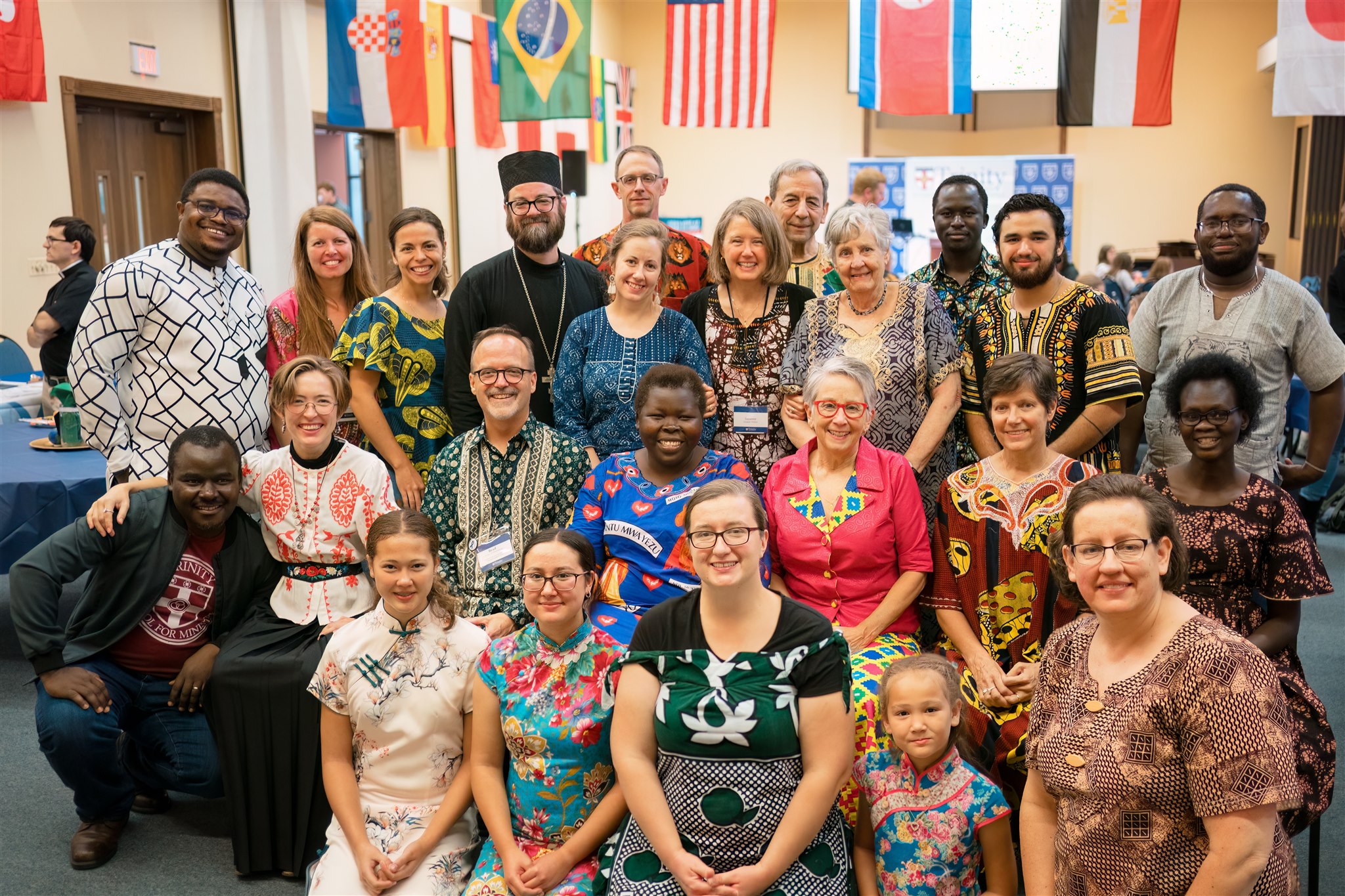 A diverse group of smiling people in colorful, traditional attire pose for a photo indoors. Multiple international flags hang in the background.