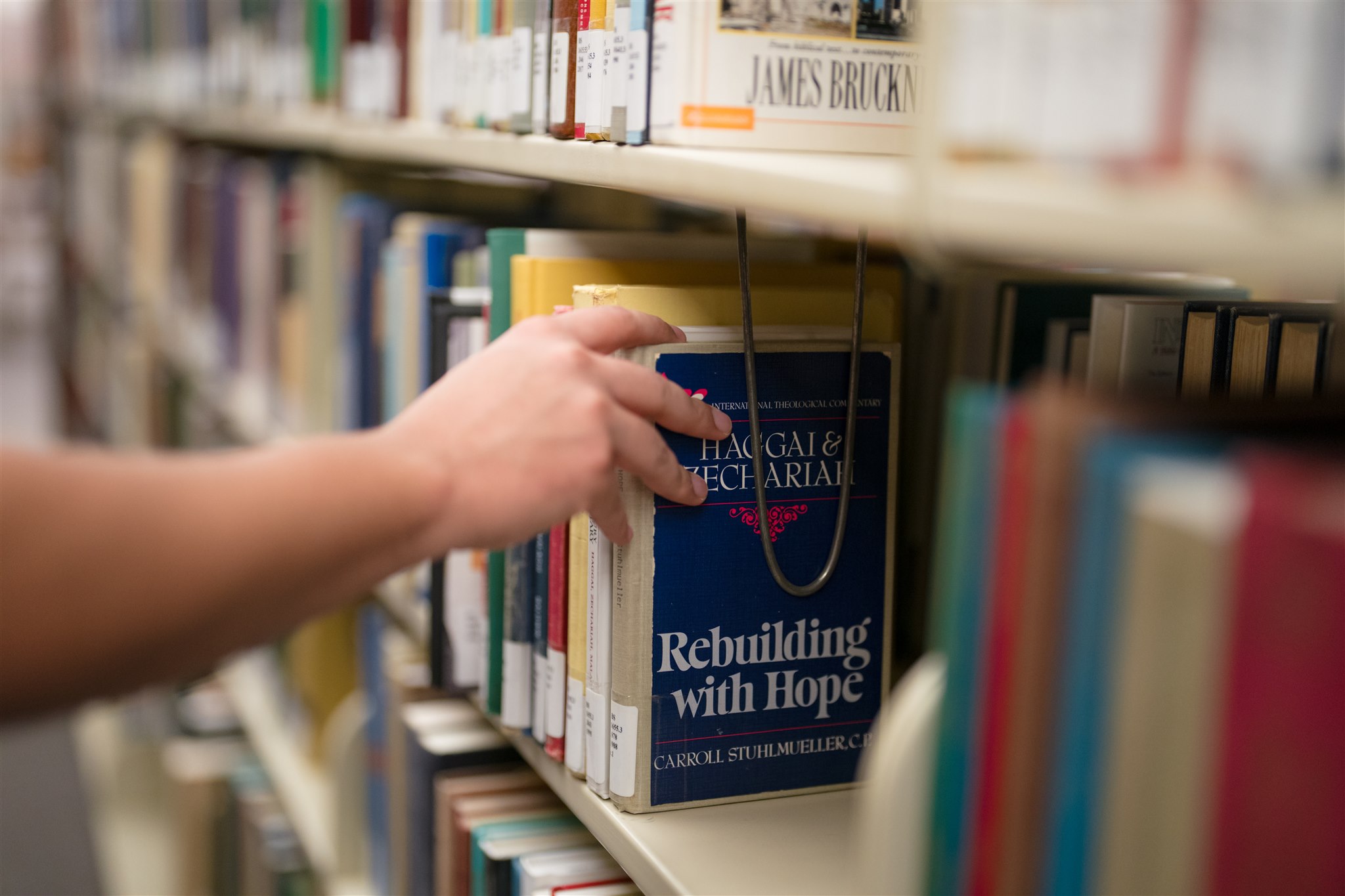 A hand reaches for a blue book titled "Rebuilding with Hope" on a library shelf, surrounded by colorful books. The scene conveys curiosity and learning.