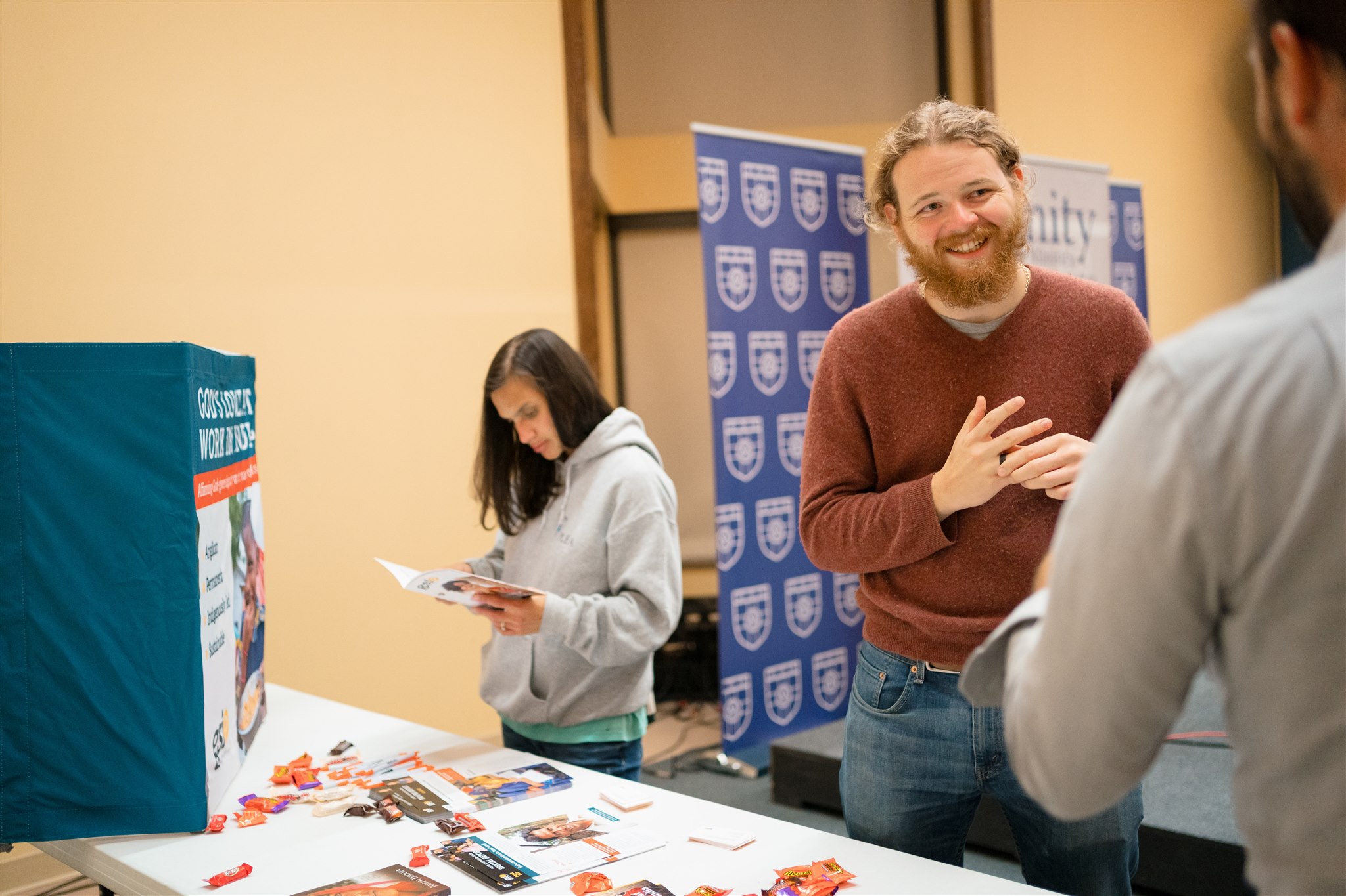 A smiling man with a beard engages with a visitor at an informational booth, featuring brochures. A woman in a hoodie reads a pamphlet nearby.