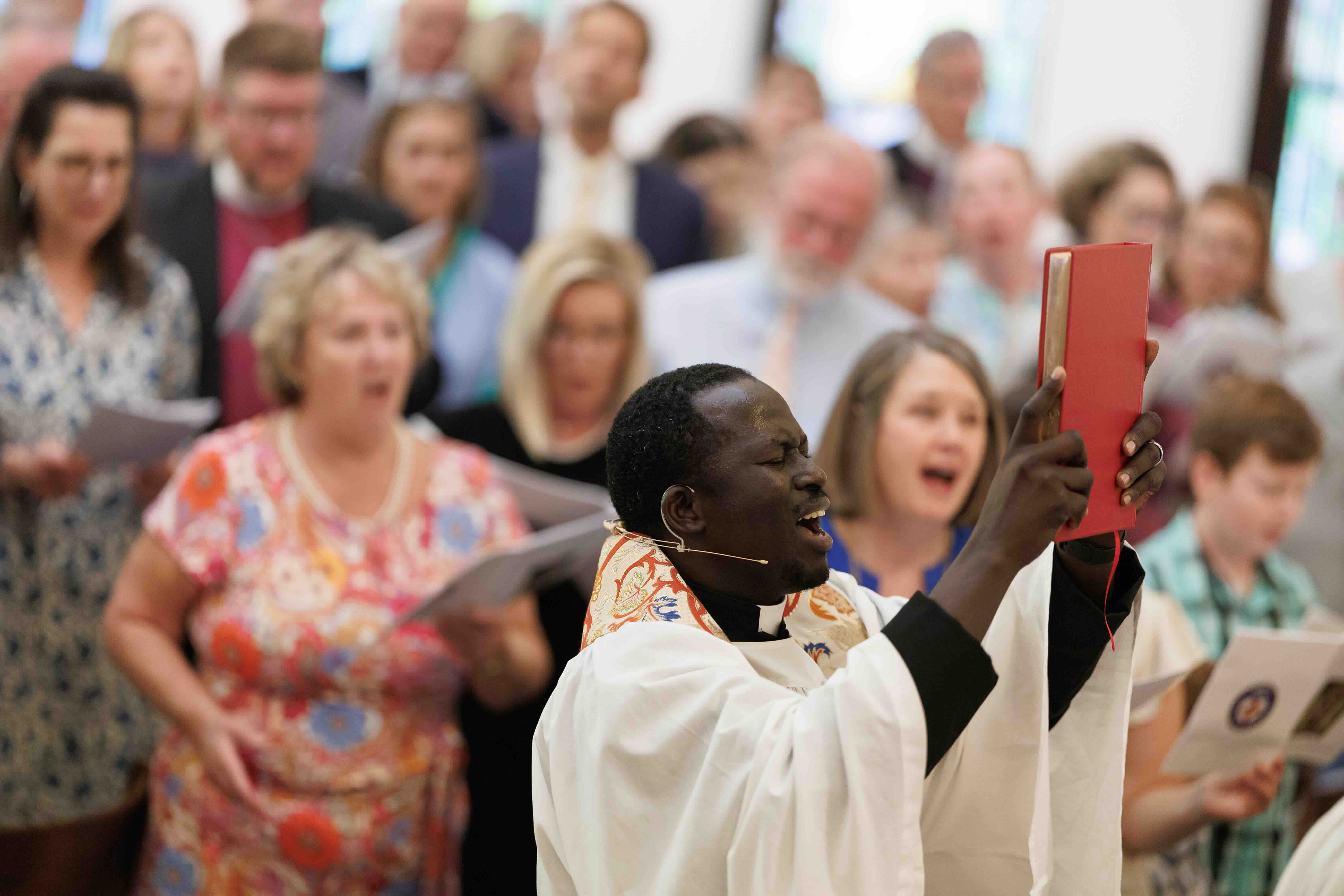 A priest in white robes holds up a red book, leading a choir in song during a church service. The group appears engaged and focused, conveying a sense of community and spirituality.