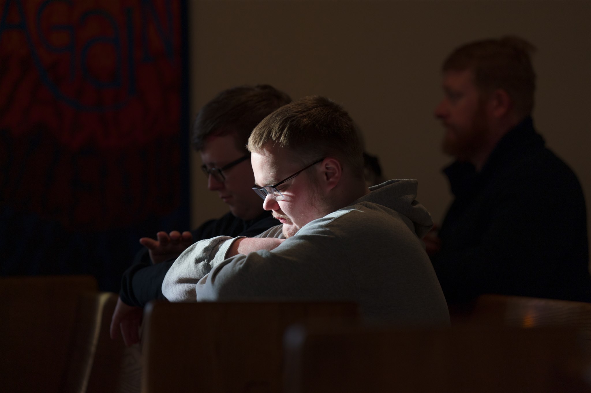 A man in glasses, wearing a gray hoodie, is seated in a dimly lit room, illuminated by soft light. He appears focused and contemplative.