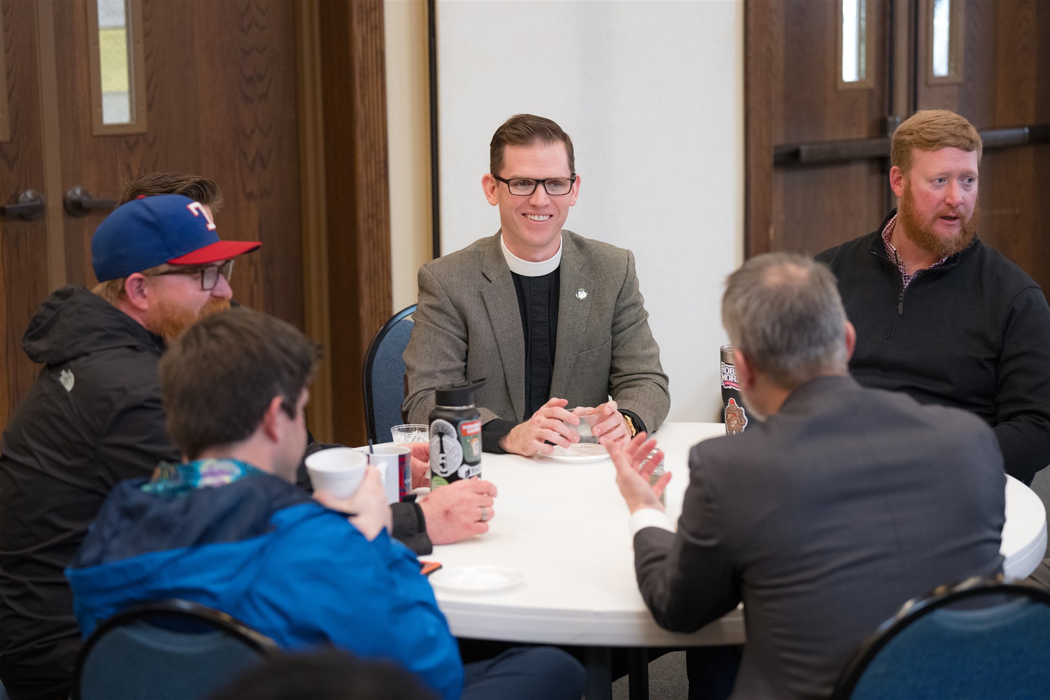 Five men are seated around a circular table in a casual meeting. They are engaged in conversation, and one man is smiling. The setting is informal.
