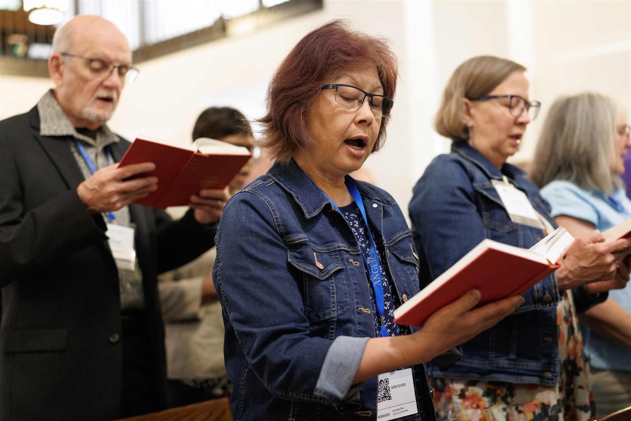 A group of people in casual attire sing from red hymnals in a bright, serene church setting, conveying a sense of community and devotion.