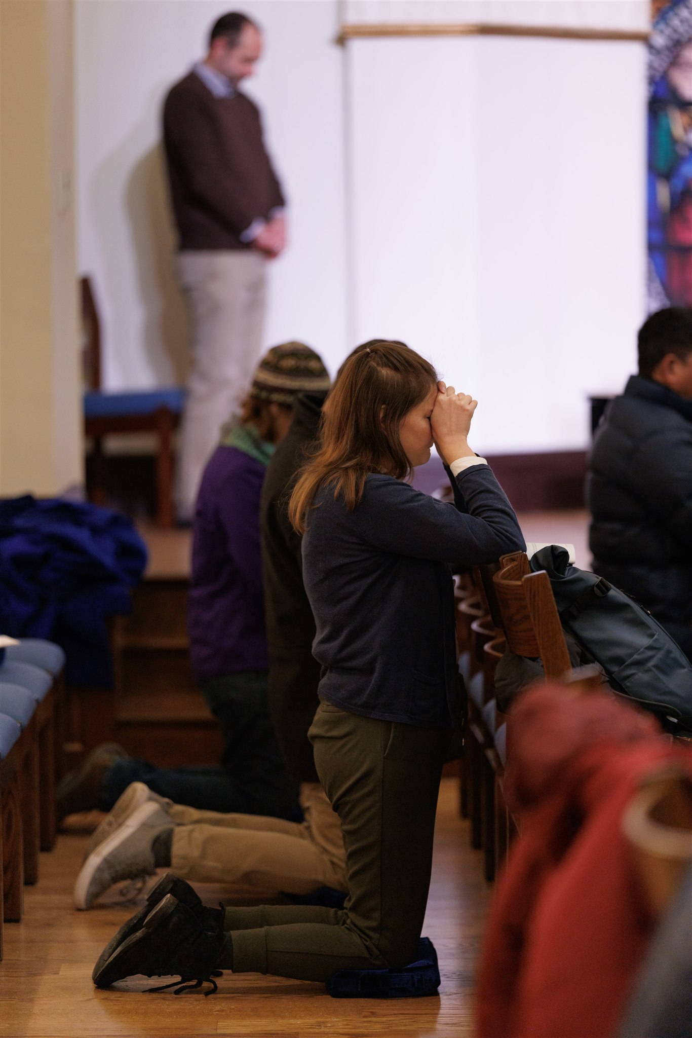 People kneeling in prayer at church pews, conveying solemnity and devotion. Background shows a standing man, blurred, adding depth to the scene.