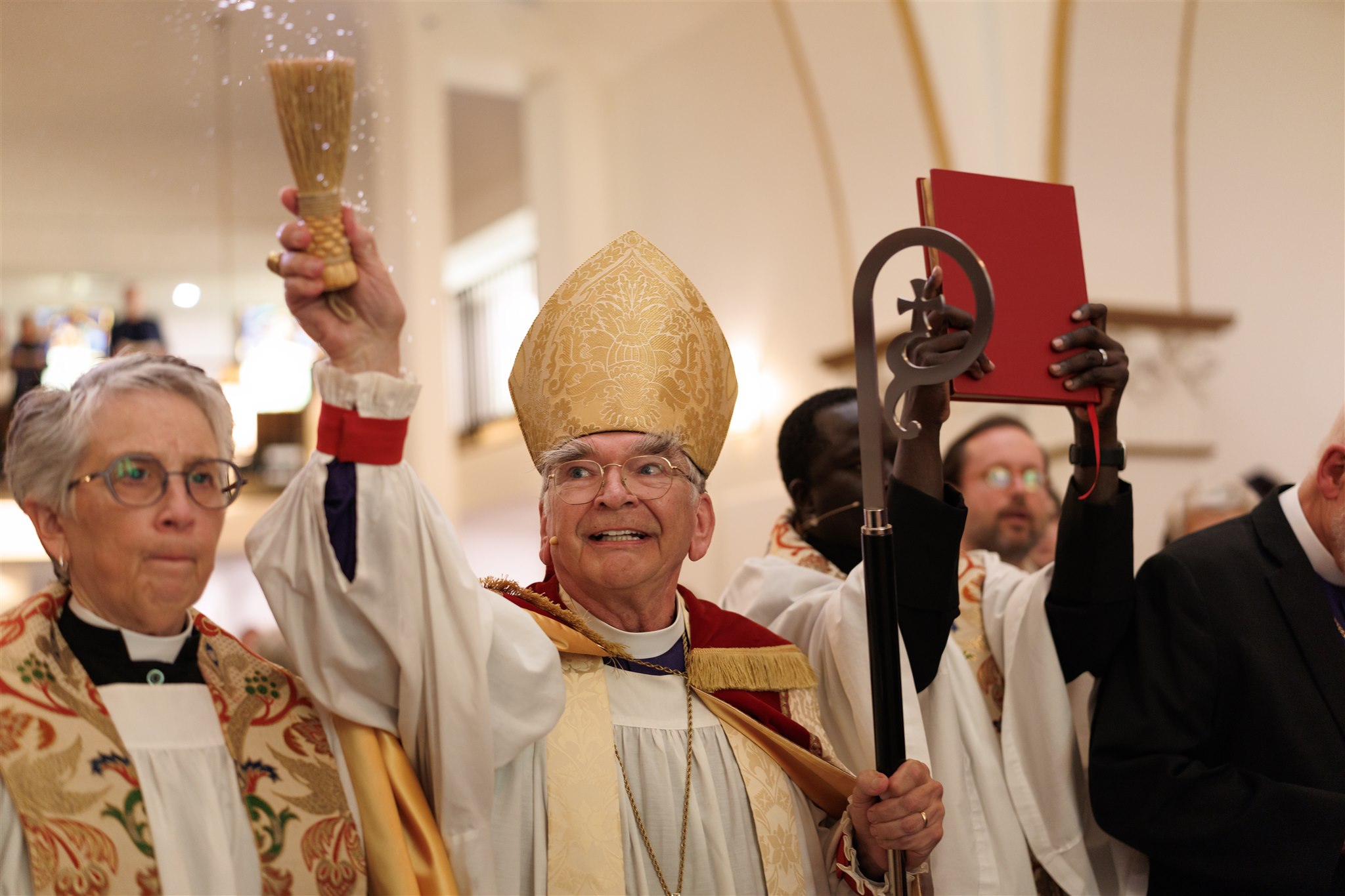 A religious leader wearing a gold mitre sprinkles holy water, smiling joyfully. Accompanied by clergy in robes, the scene conveys celebration and blessing.