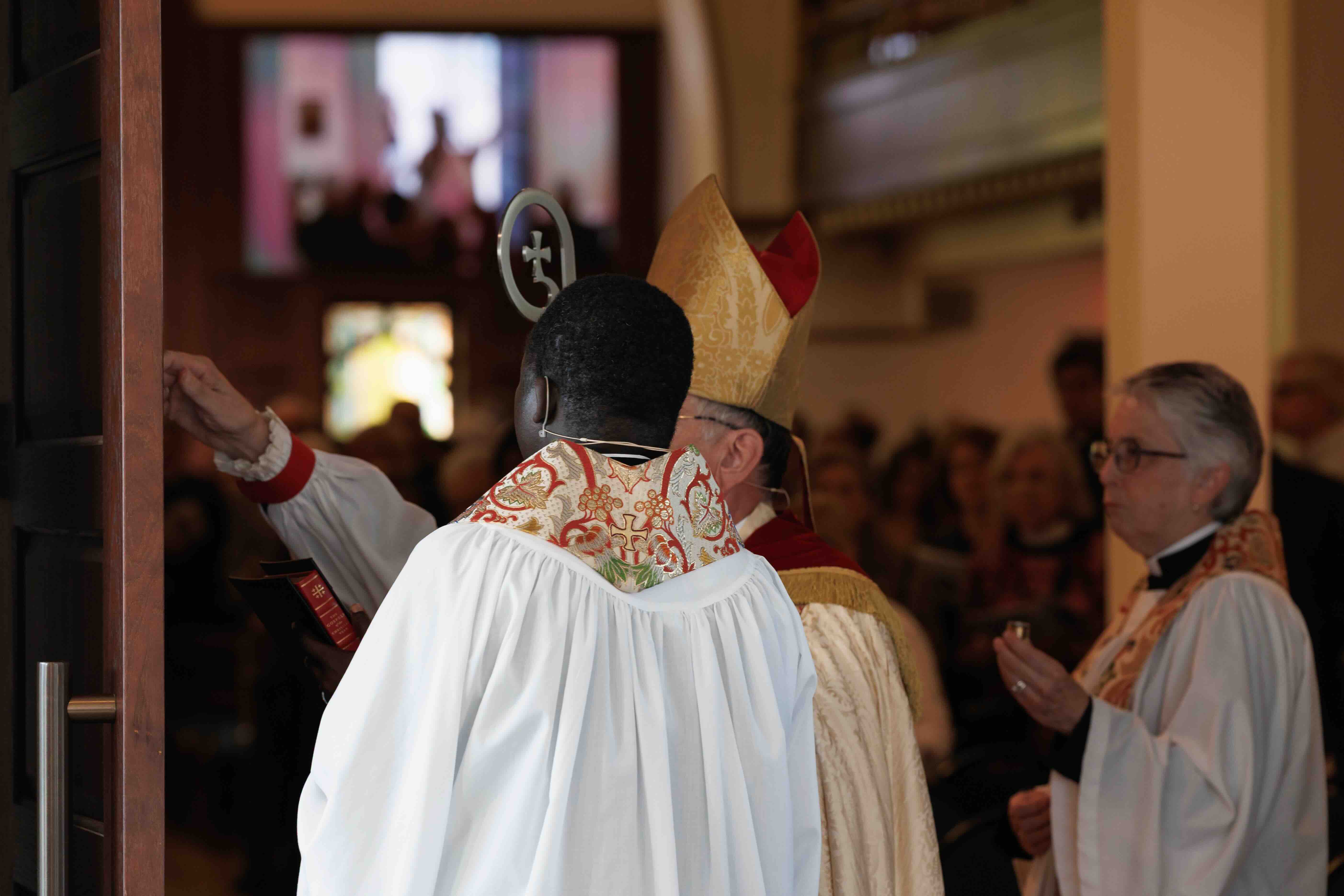 A religious ceremony with a bishop in gold and red robes, holding a staff, faces a church interior. Attendees in white clergy robes observe solemnly.