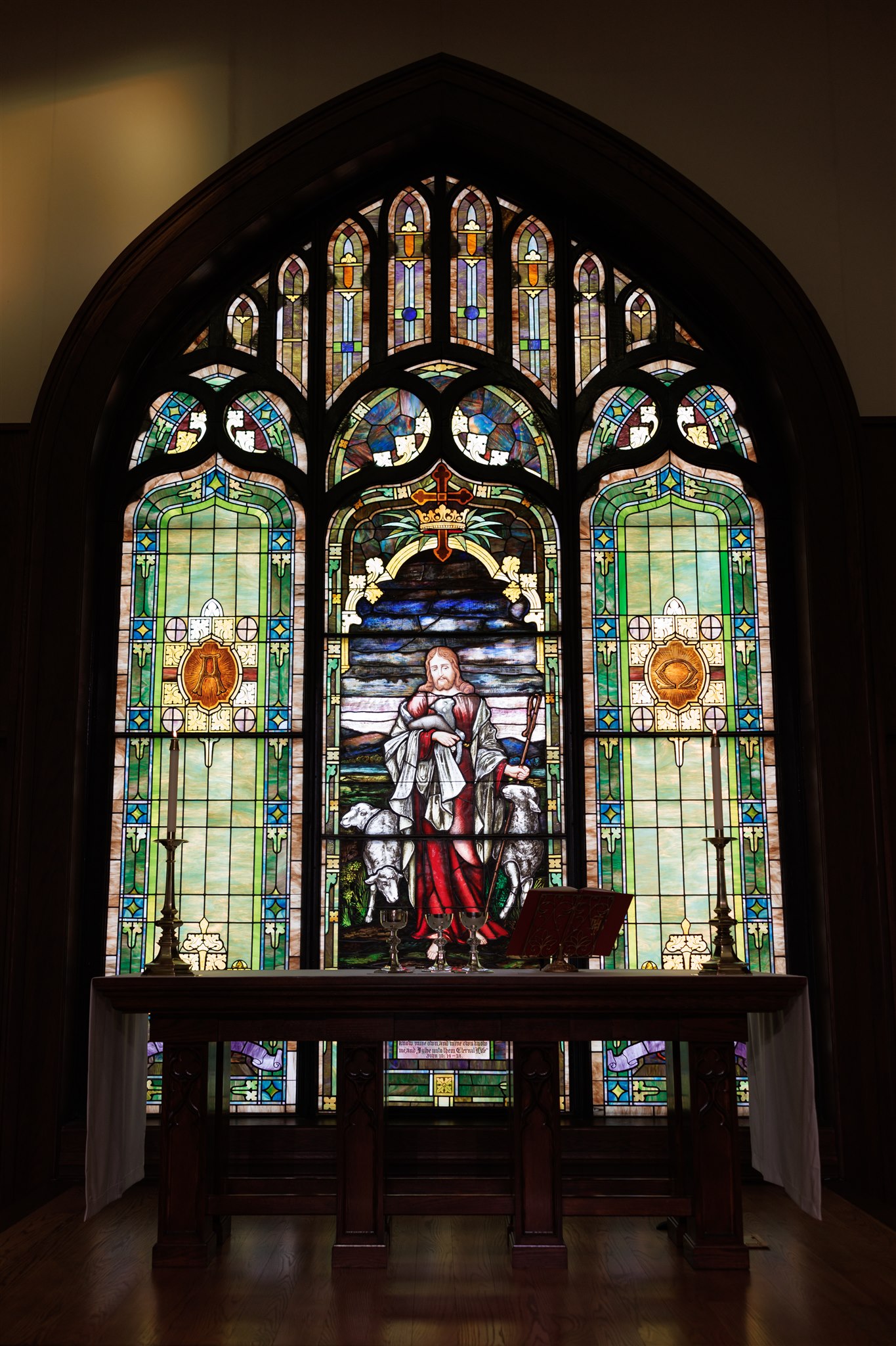 Stained glass window depicting a serene religious scene with a central figure holding a staff, flanked by sheep. Rich colors enhance the sacred tone.