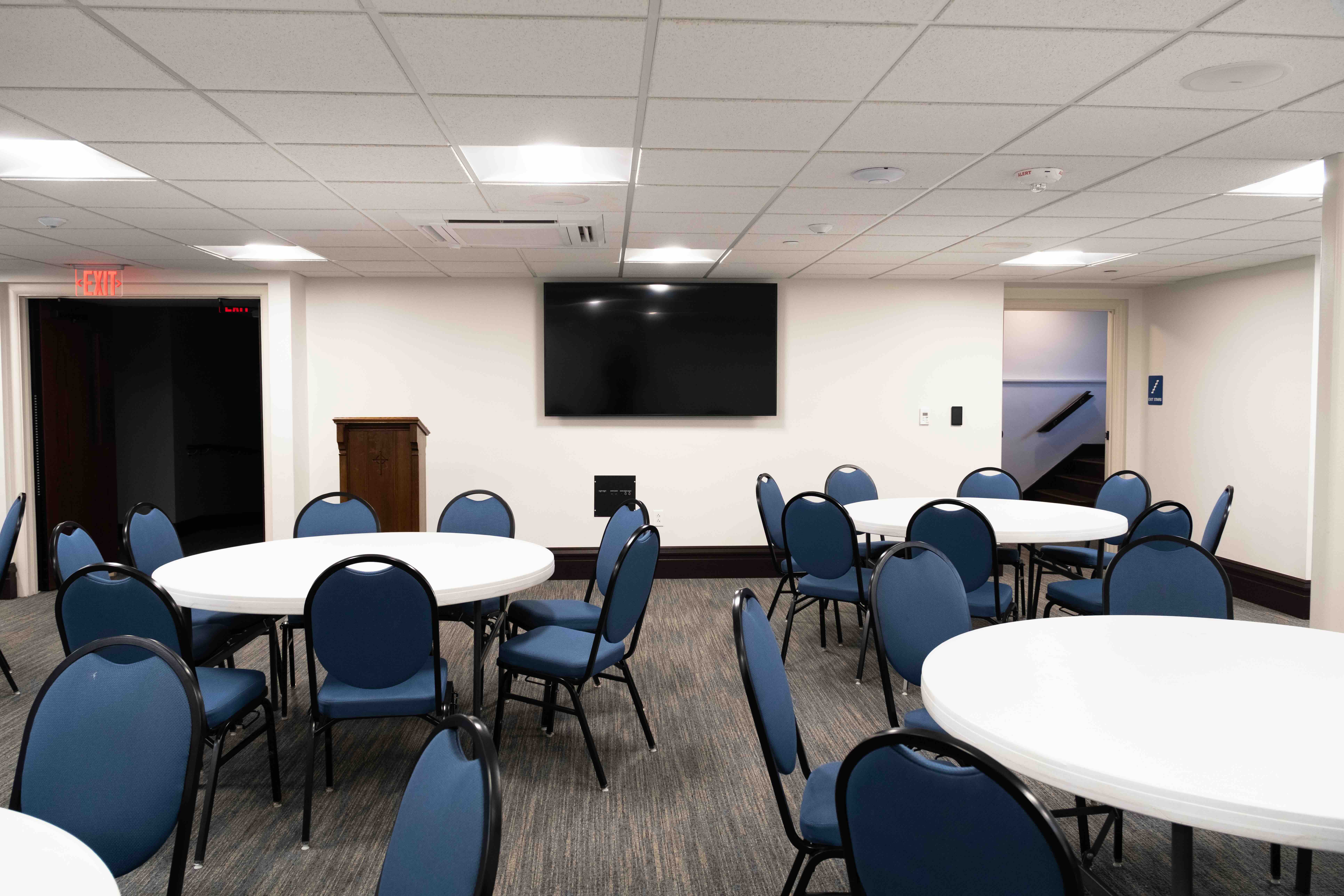 Spacious conference room with blue chairs and round tables arranged neatly. A podium and large TV screen are at the front. Neutral colors create a professional atmosphere.