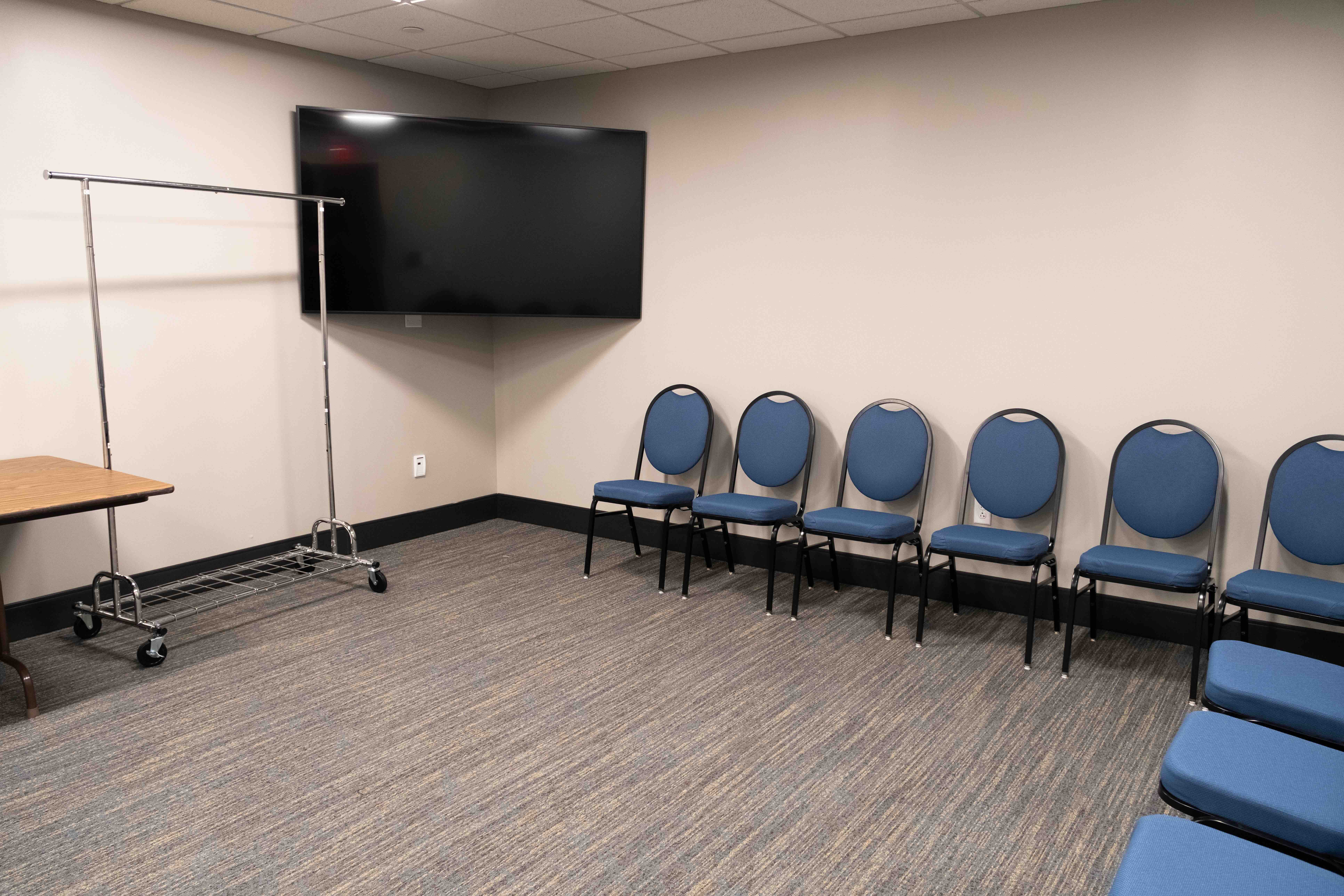 Meeting room with blue chairs lined against a beige wall, a large flat-screen TV, a clothes rack, and a folding table. The setting is simple and tidy.