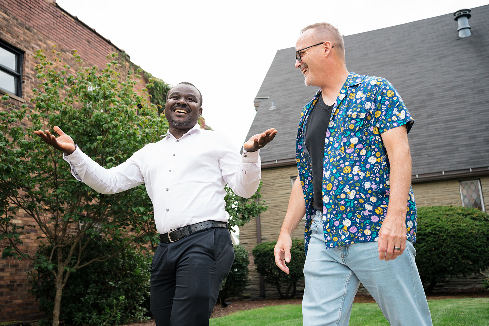 Two men happily walk outside, talking and smiling. One wears a white shirt; the other, a colorful floral shirt. They appear relaxed and joyful.