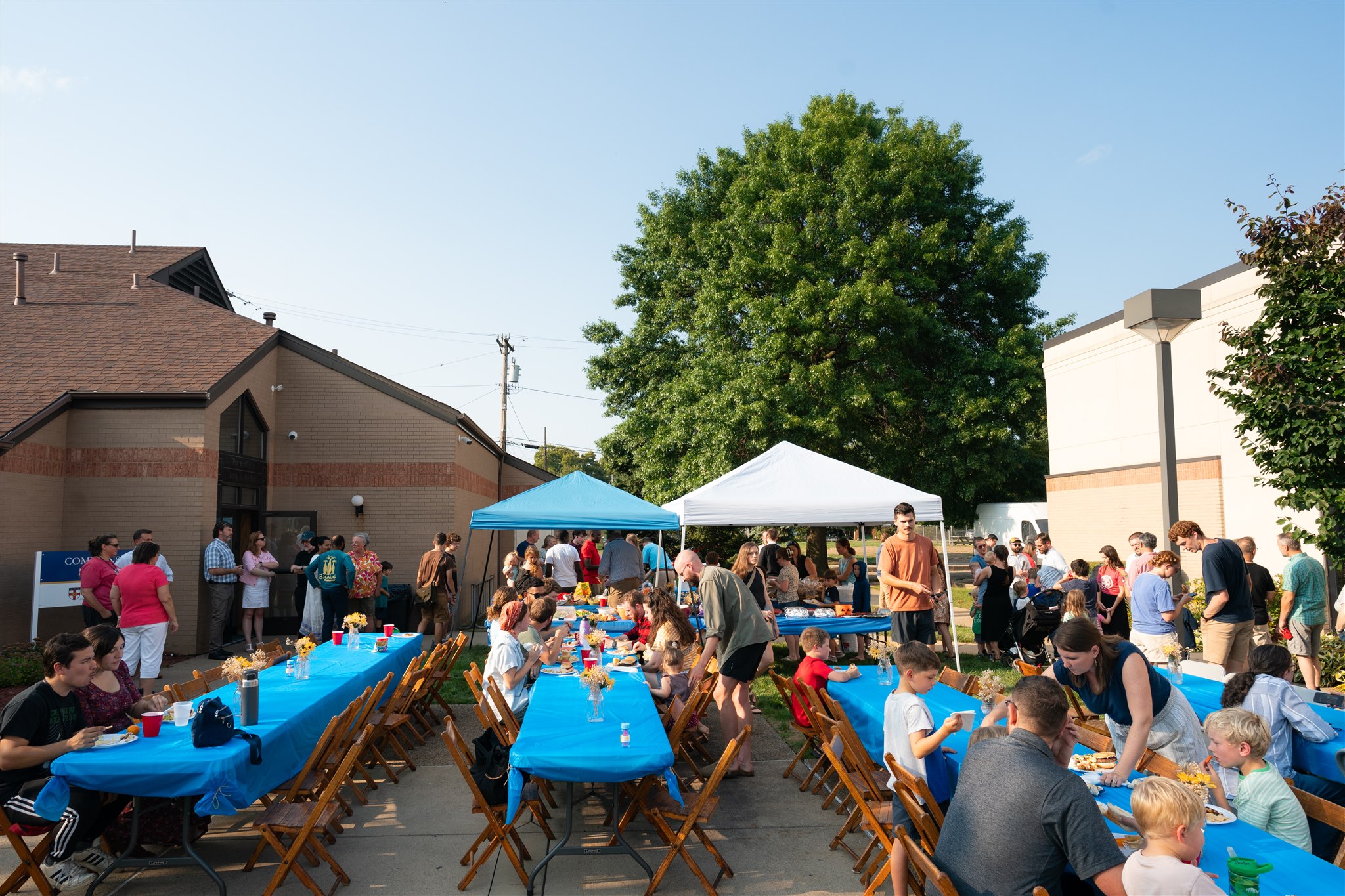 Outdoor gathering with people sitting at long tables covered with blue tablecloths. There's a festive atmosphere. Trees and a building are in the background.