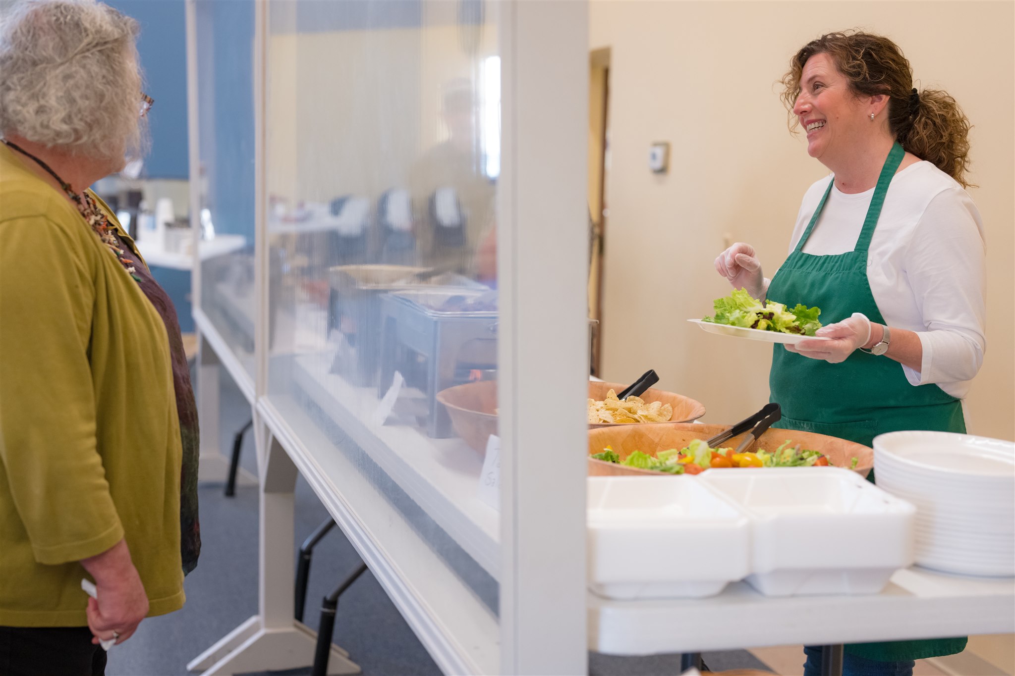 A woman in a green apron serves food with a smile at a cafeteria counter. Another woman with gray hair stands across, creating a warm, friendly atmosphere.