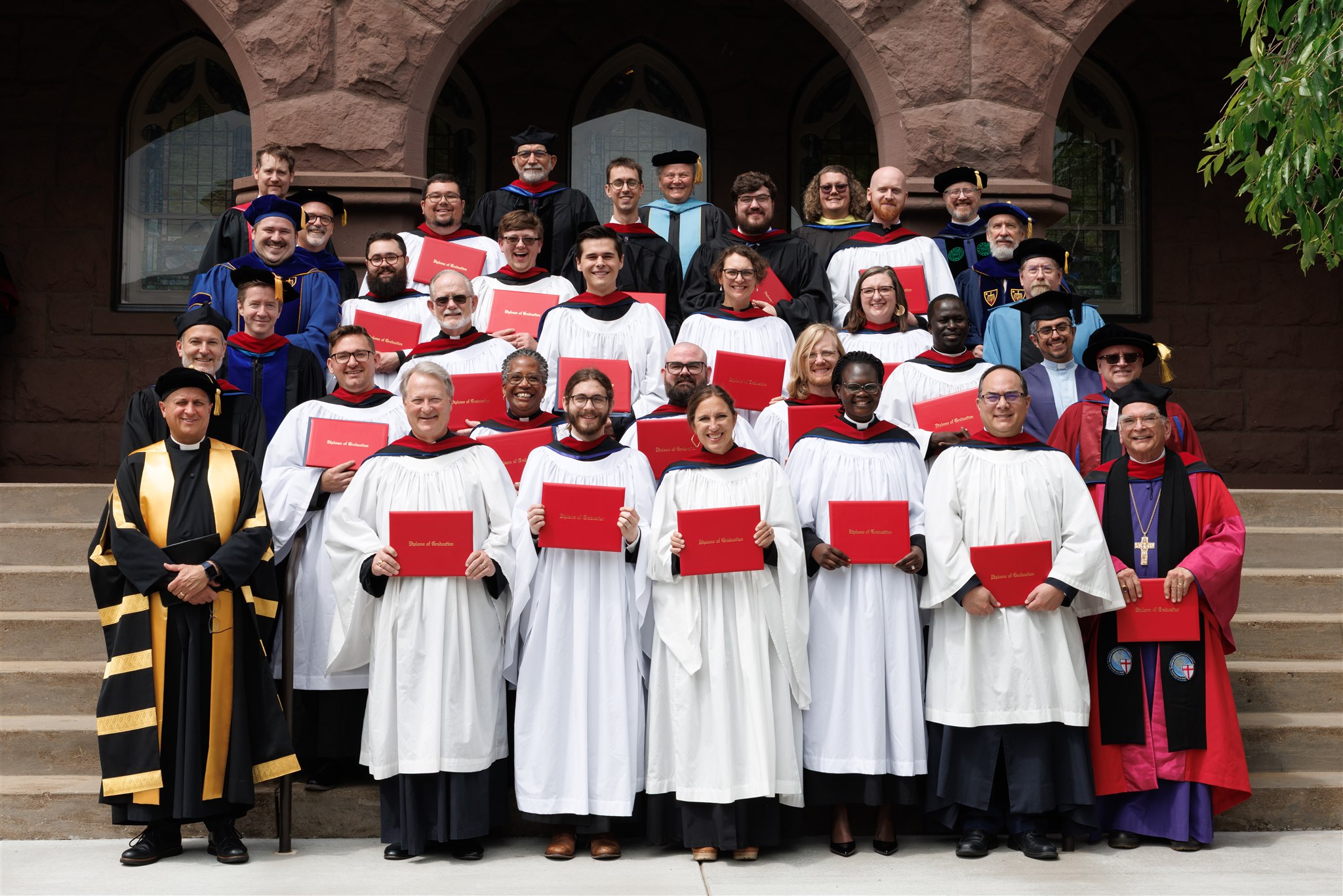 A large group of people in graduation gowns and academic regalia gather on steps, smiling and holding red diplomas. The mood is celebratory and proud.