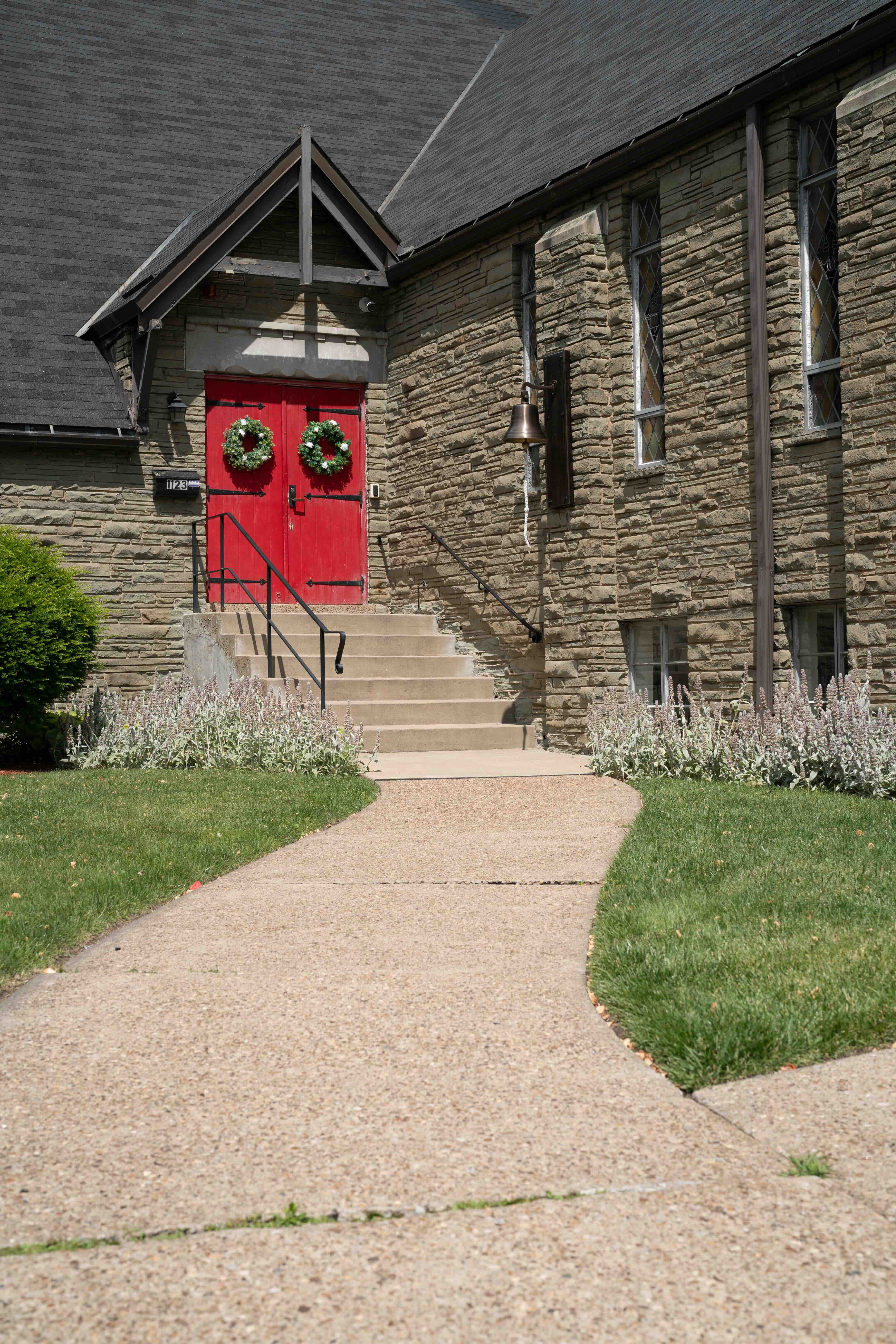 Stone building with red double doors decorated with green wreaths, steps with handrails, and a large brass bell on the wall.