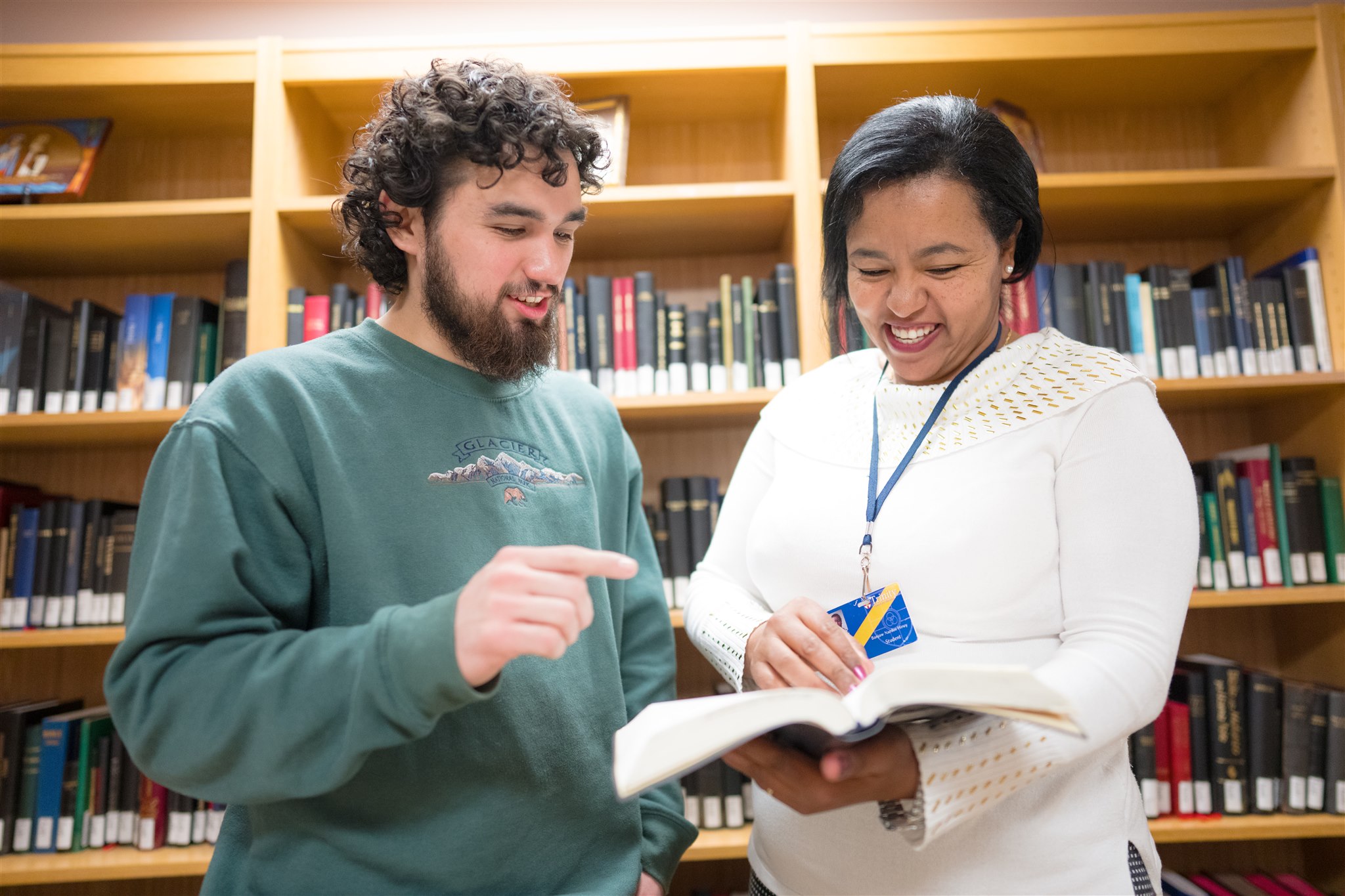 A man and woman smiling and discussing a book in a library. The man points at the book while the woman holds it. Bookshelves are in the background.