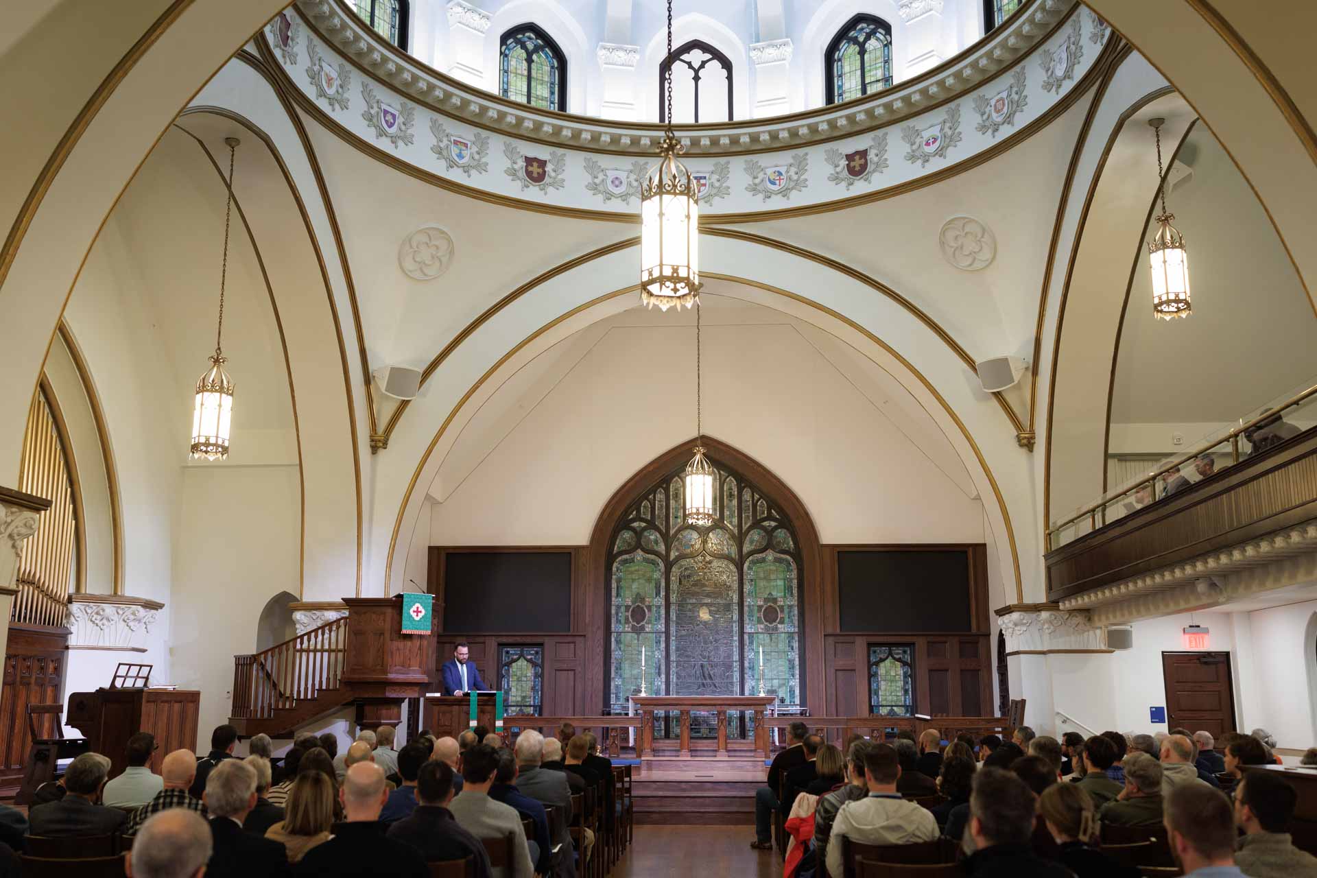 Audience seated in a church listening to a speaker at a wooden pulpit in front of a stained glass window.