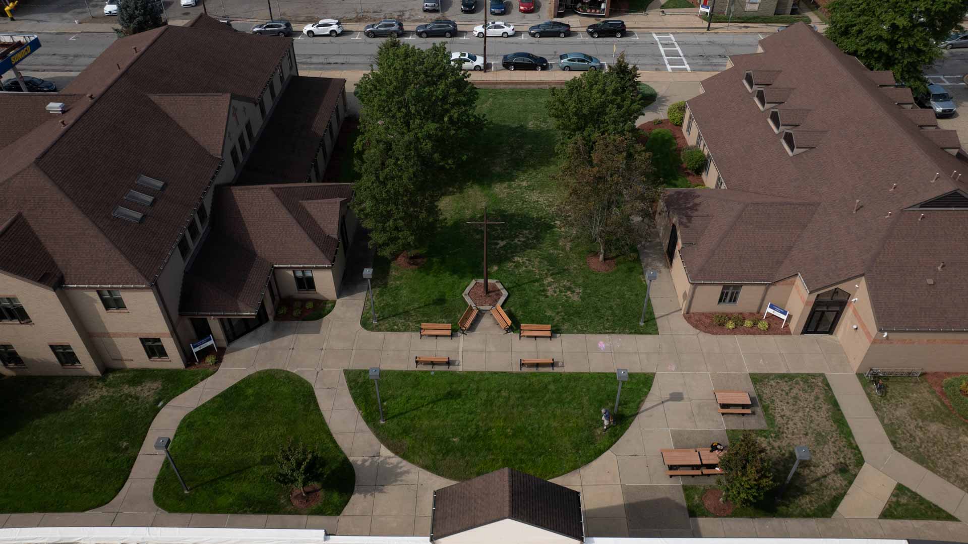 Aerial view of campus courtyard between academic buildings, with central cross, trees, benches, and walkways.