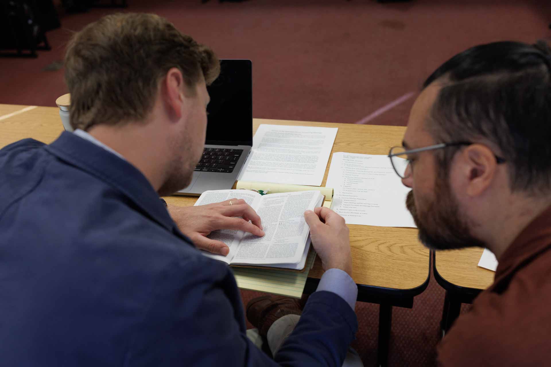 Two men seated at a table reviewing an open book with papers and a laptop in front of them.