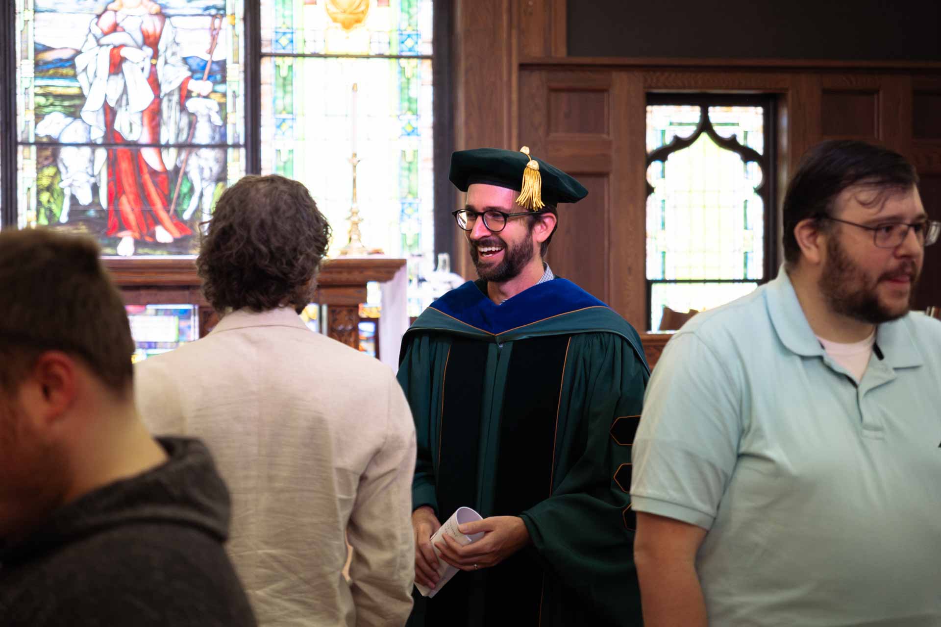 A bearded man in traditional academic regalia, including a dark green doctoral gown and tam with gold tassel, smiles warmly during a graduation ceremony in a chapel with stained glass windows visible in the background