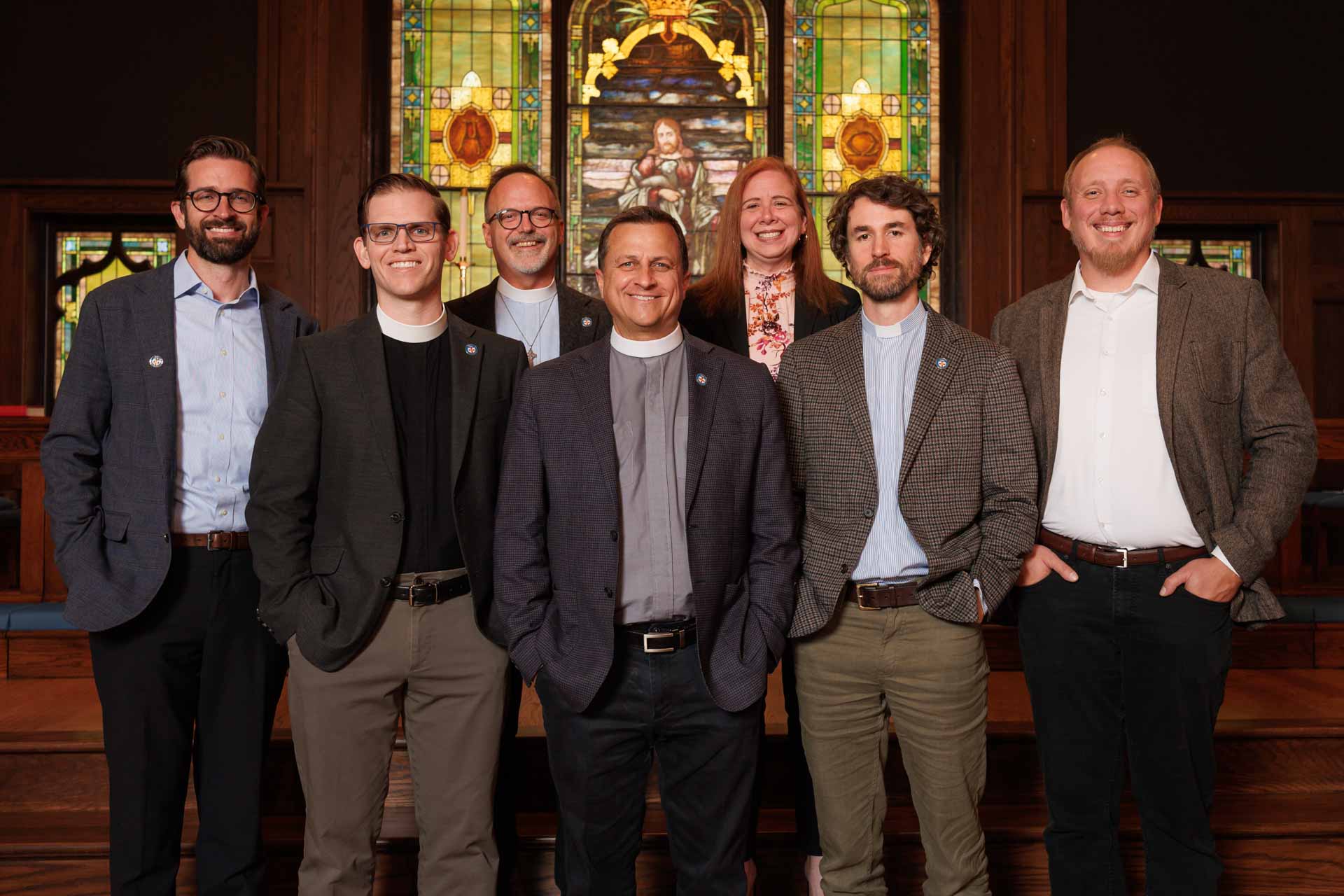 Seven people including clergy members standing in front of a stained glass window inside a church.