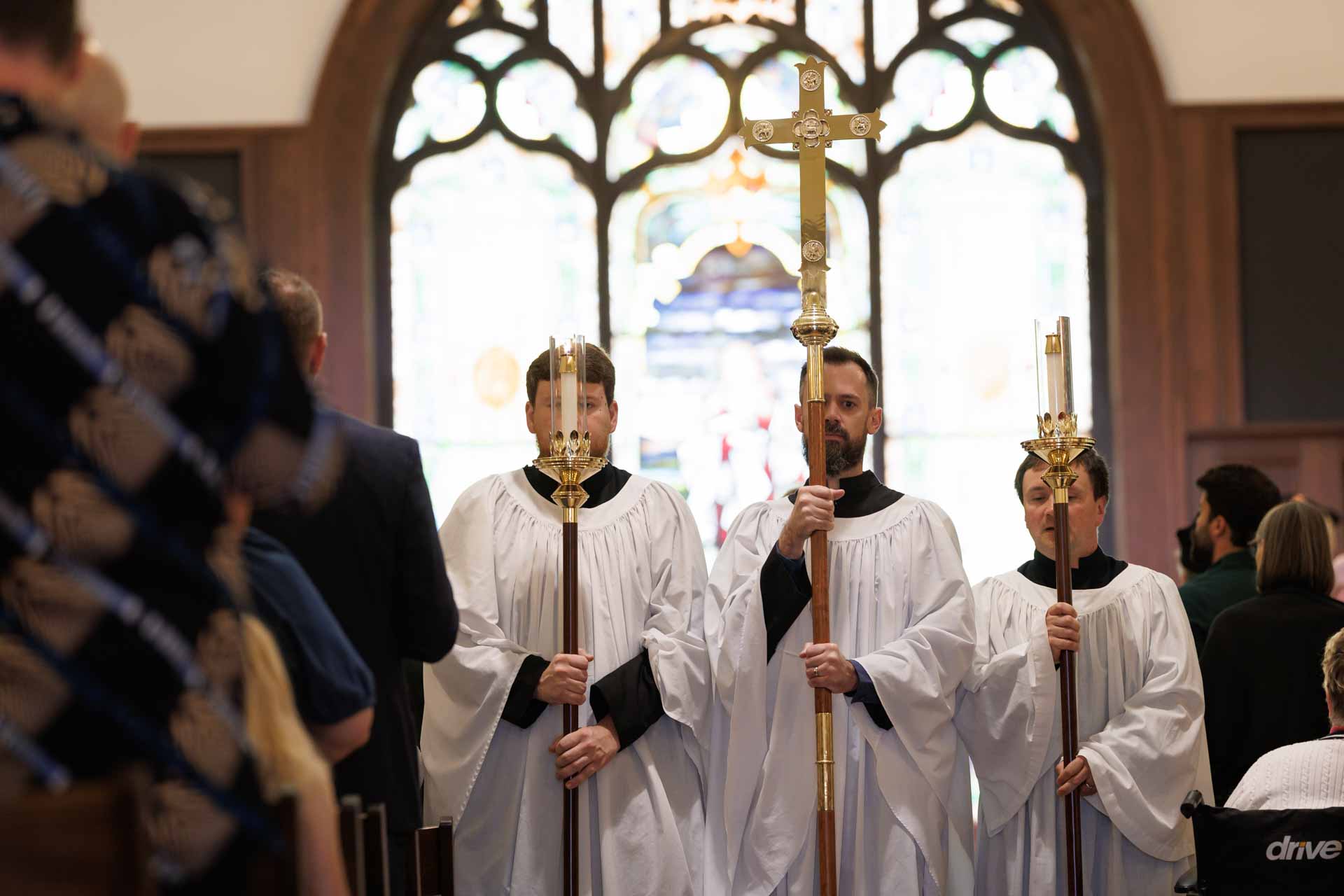 Three men in white church robes holding a cross and two candles during a religious procession inside a church.