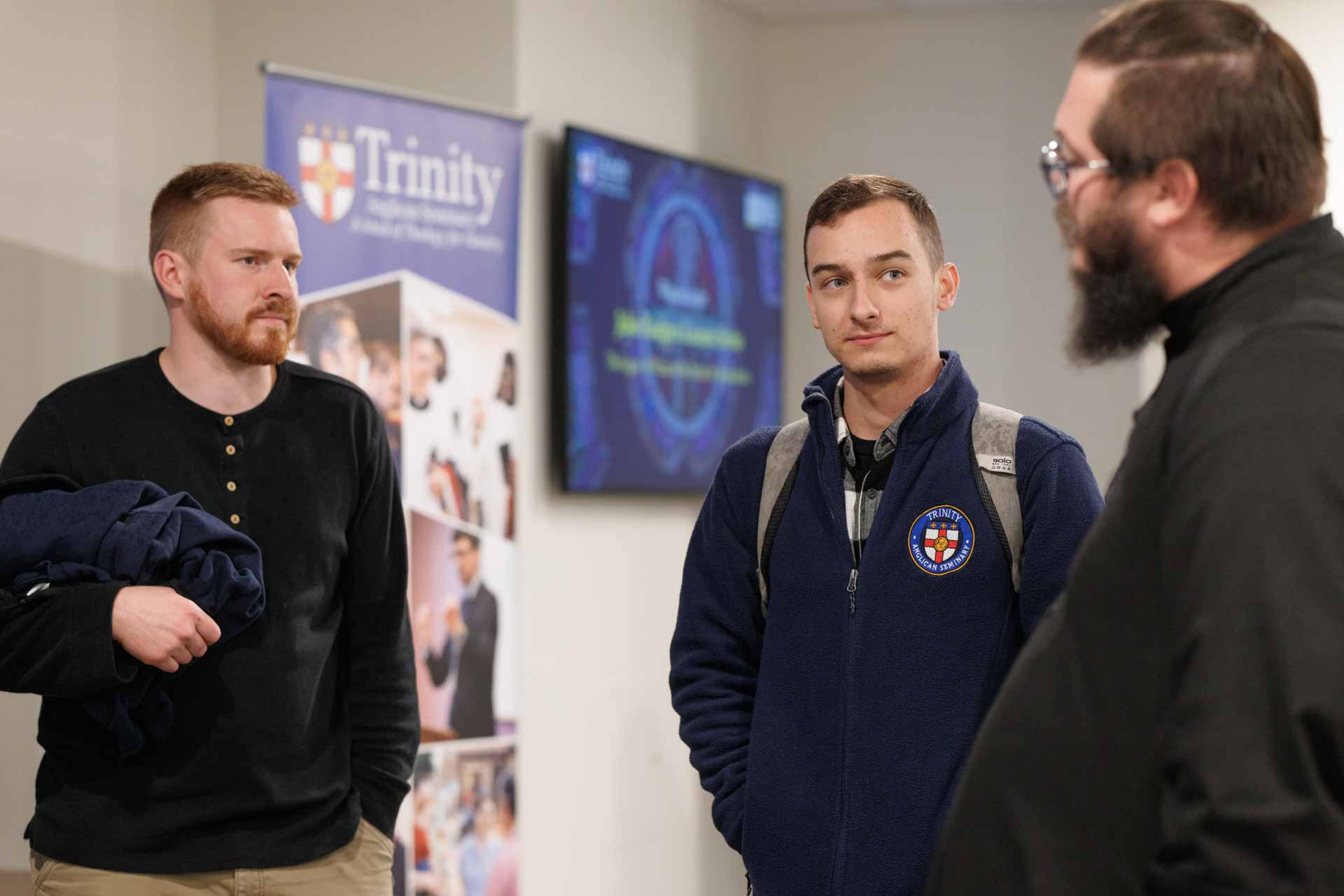 Three men engaged in conversation indoors; one wearing a Trinity Anglican Seminary jacket and a backpack.