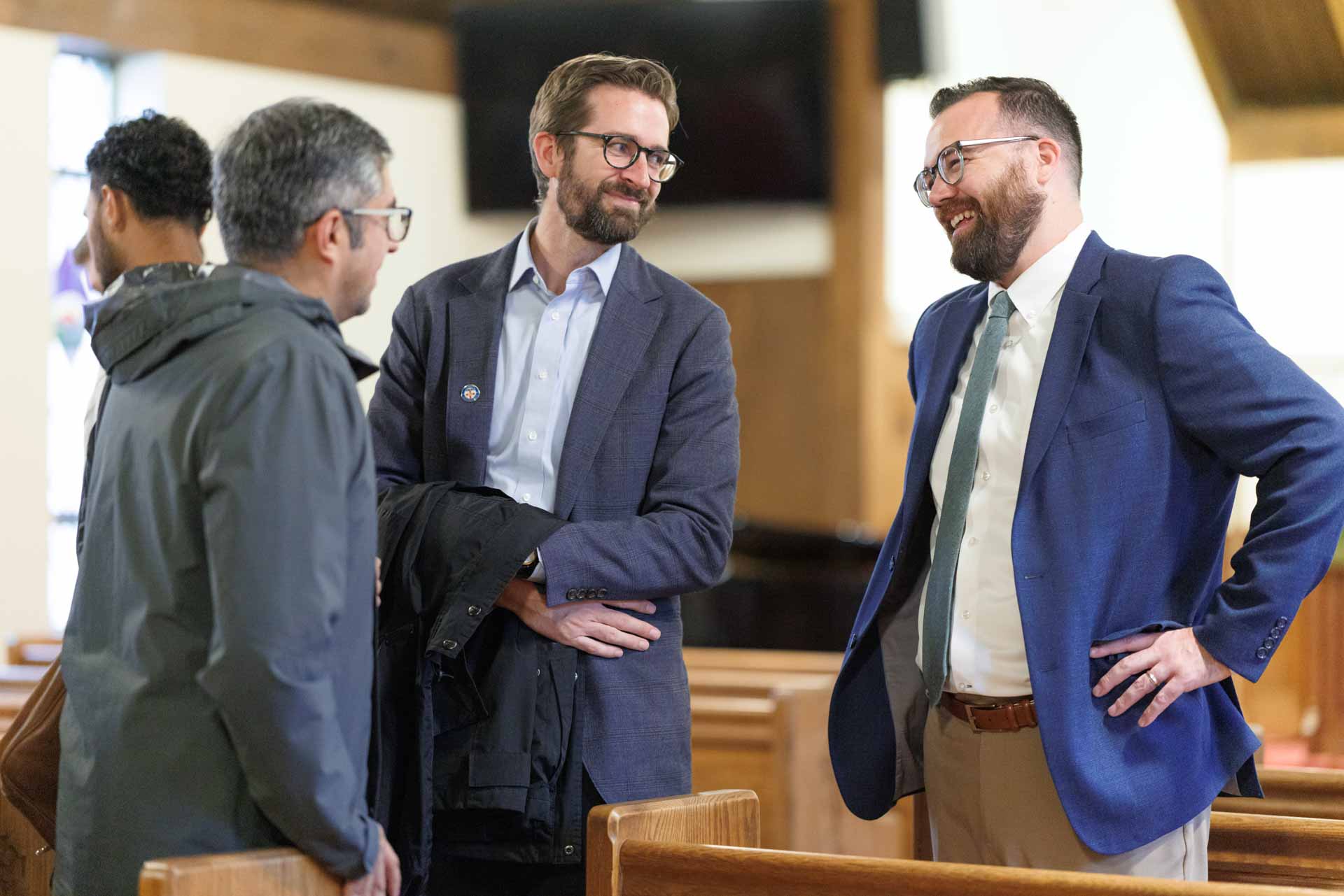 Four men engaged in conversation inside a wood-paneled room with benches, two men in suits smiling and one man wearing a gray jacket.