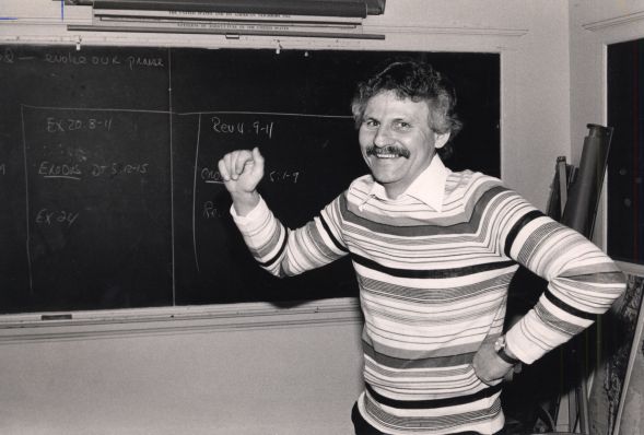Smiling man with a mustache wearing a striped sweater, standing in front of a blackboard with handwritten notes.