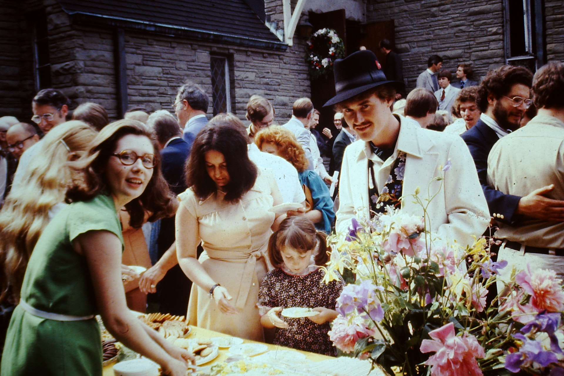 People gathered outdoors at a social event around a table with snacks and flowers, including a man in a white suit with a black hat and a young girl holding a plate.