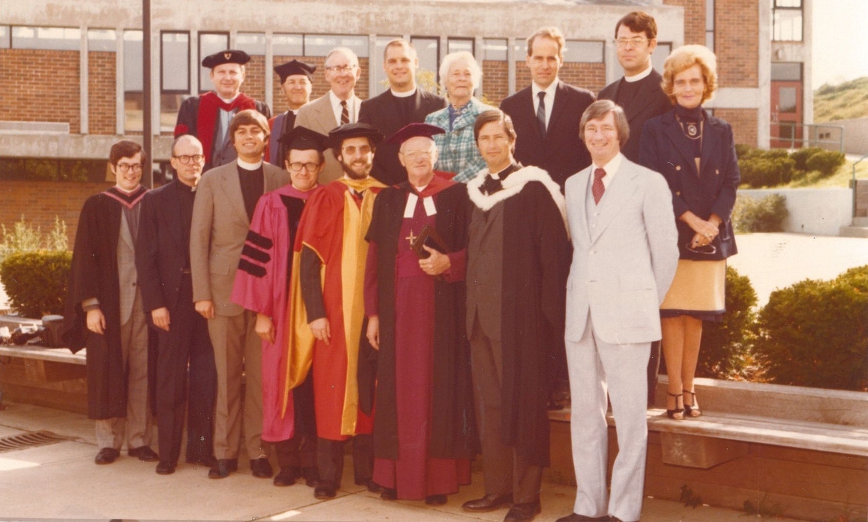 Group portrait of 17 faculty members in academic regalia and formal wear standing outside a brick building.