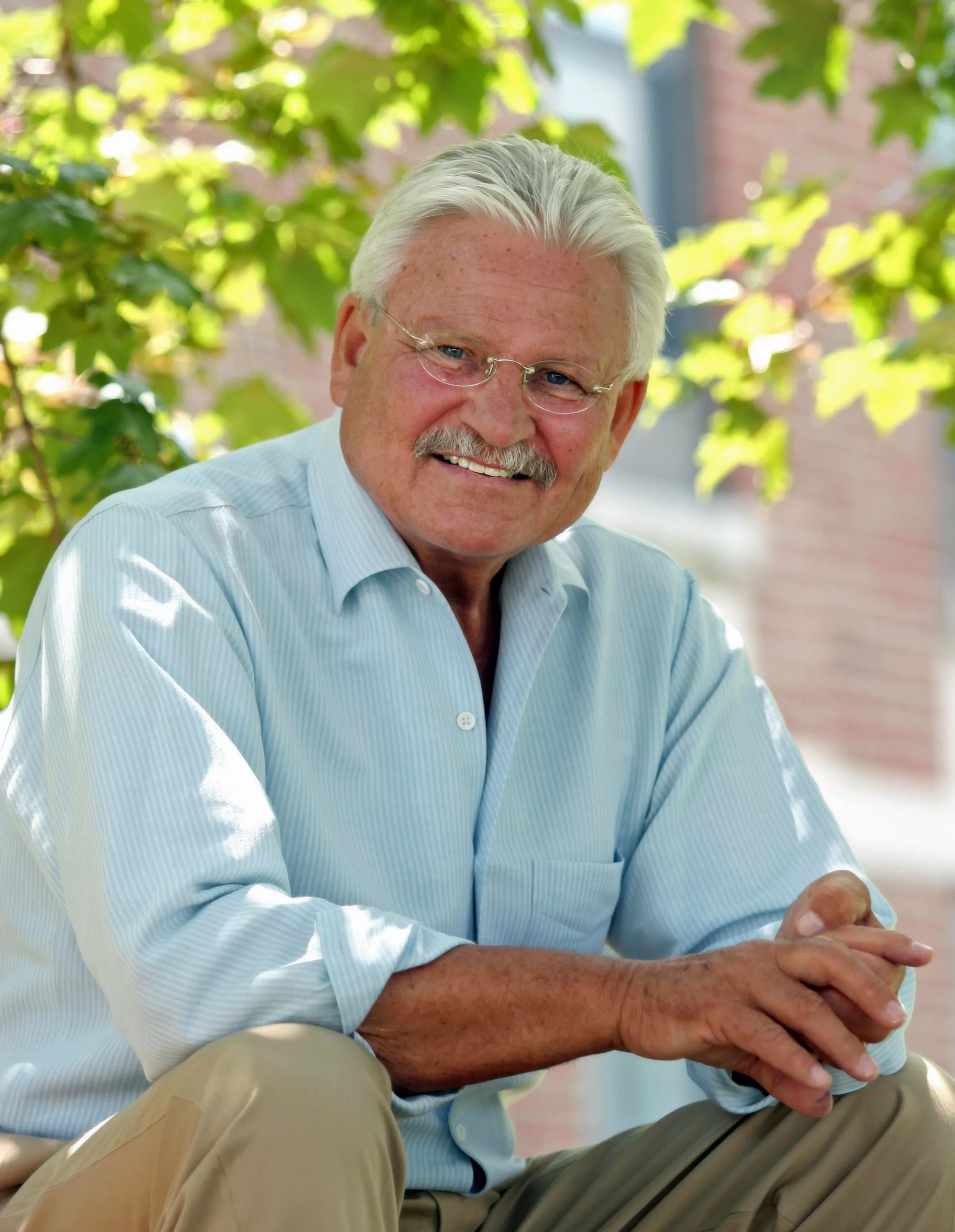 Older man with white hair, mustache, and glasses smiling outdoors with green leaves and brick wall background.