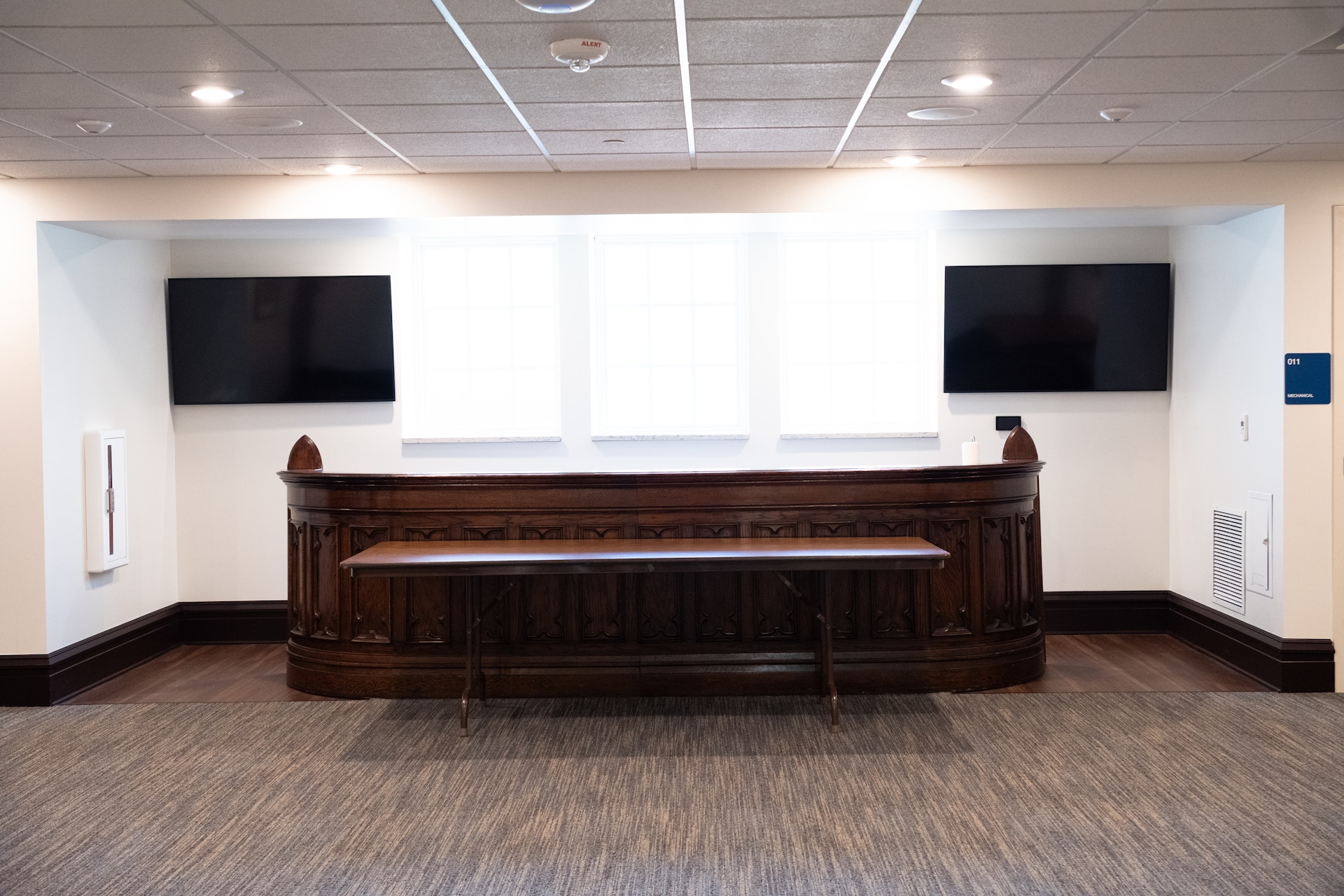 Room with a dark wooden counter in front of three large windows, two wall-mounted flat-screen TVs, and a wooden folding table in front.