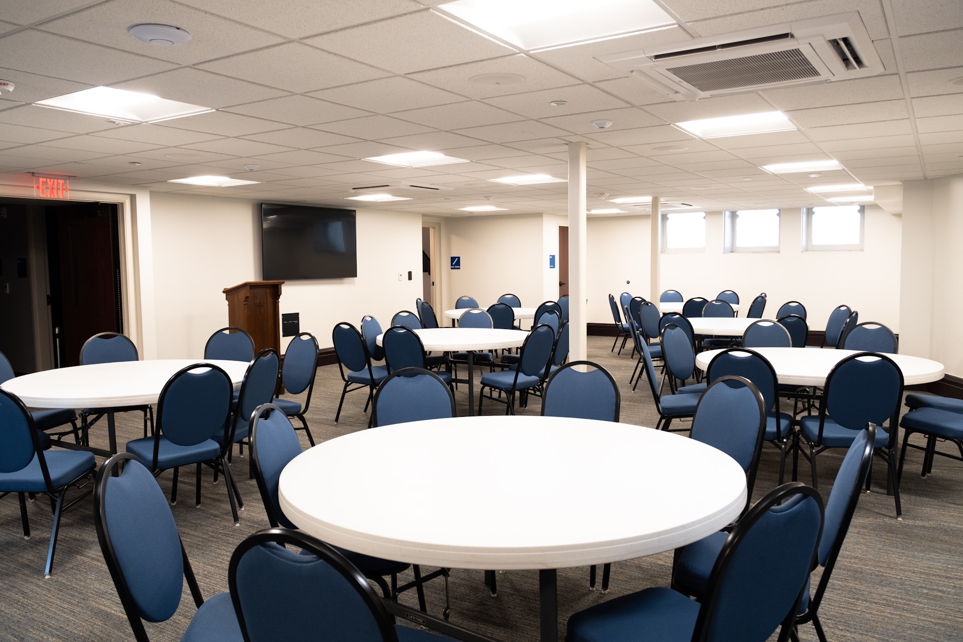 Room with multiple round white tables surrounded by blue chairs, a wall-mounted TV, and a wooden podium under bright ceiling lights.