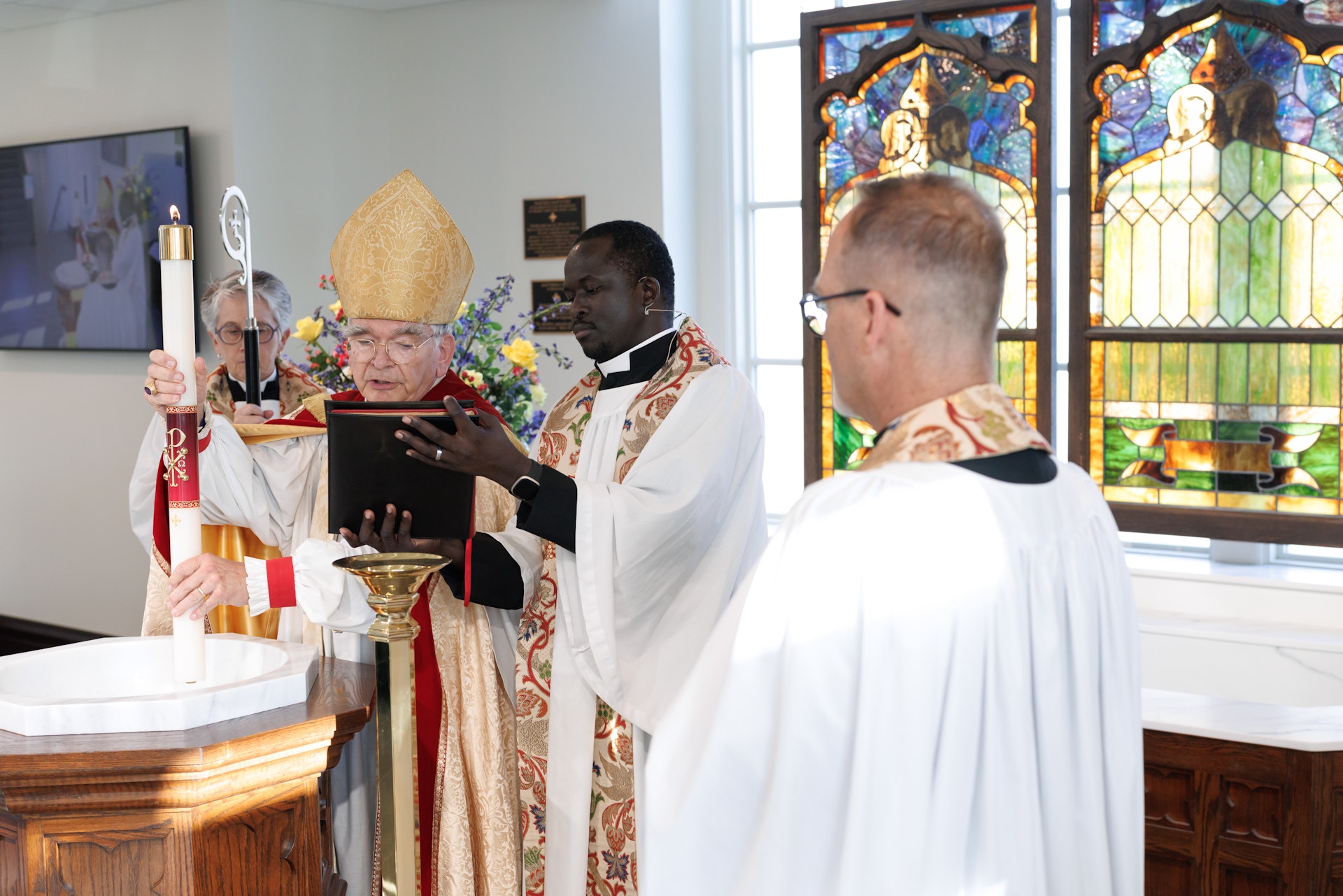 Clergy members in liturgical robes conduct a religious ceremony around a wooden baptismal font with stained glass windows in the background.