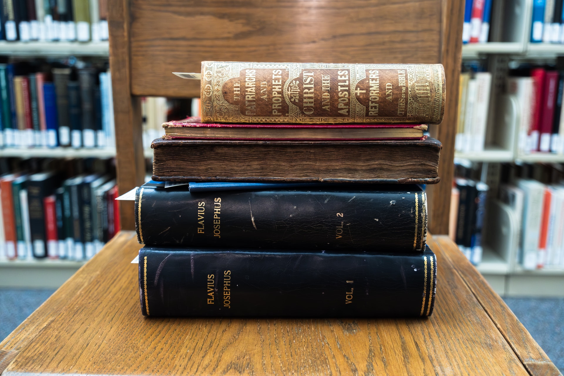 Stack of old books on a wooden table in a library, with shelves of books blurred in the background.