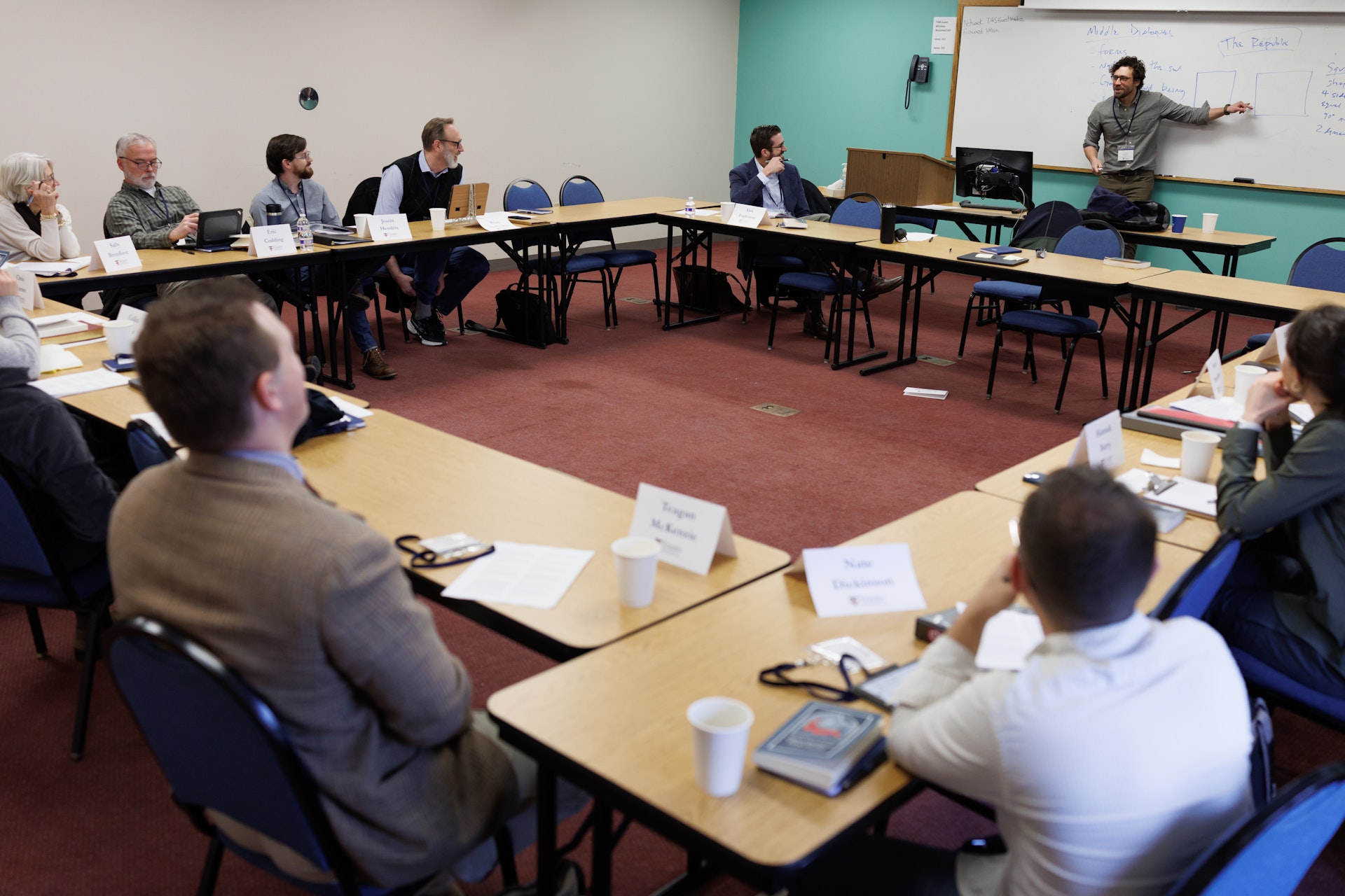 Group of adults seated around U-shaped tables attentively listening to a presenter pointing at notes on a whiteboard in a classroom.