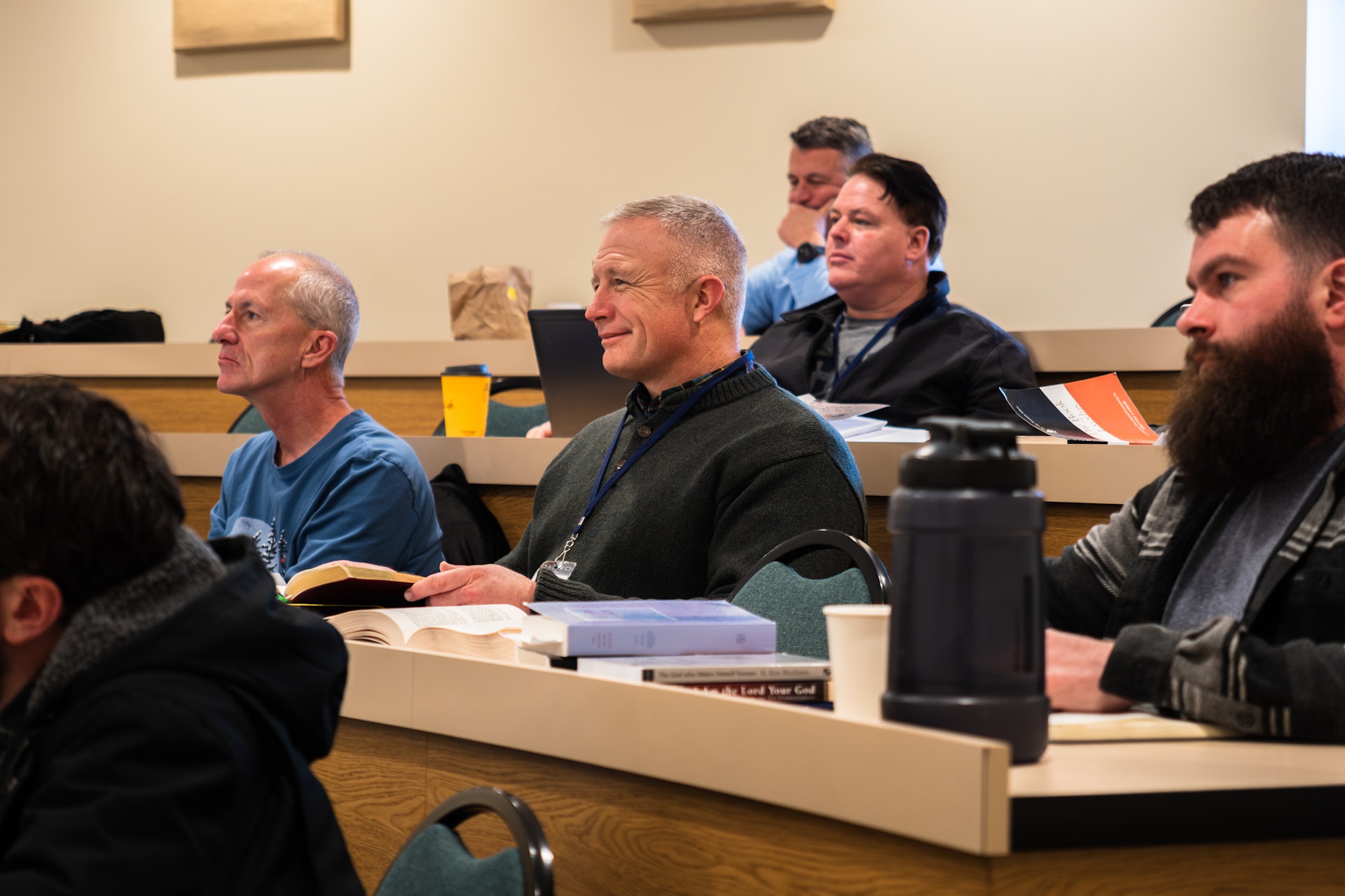 Men attentively sitting in a classroom with open books, papers, and personal items on desks.