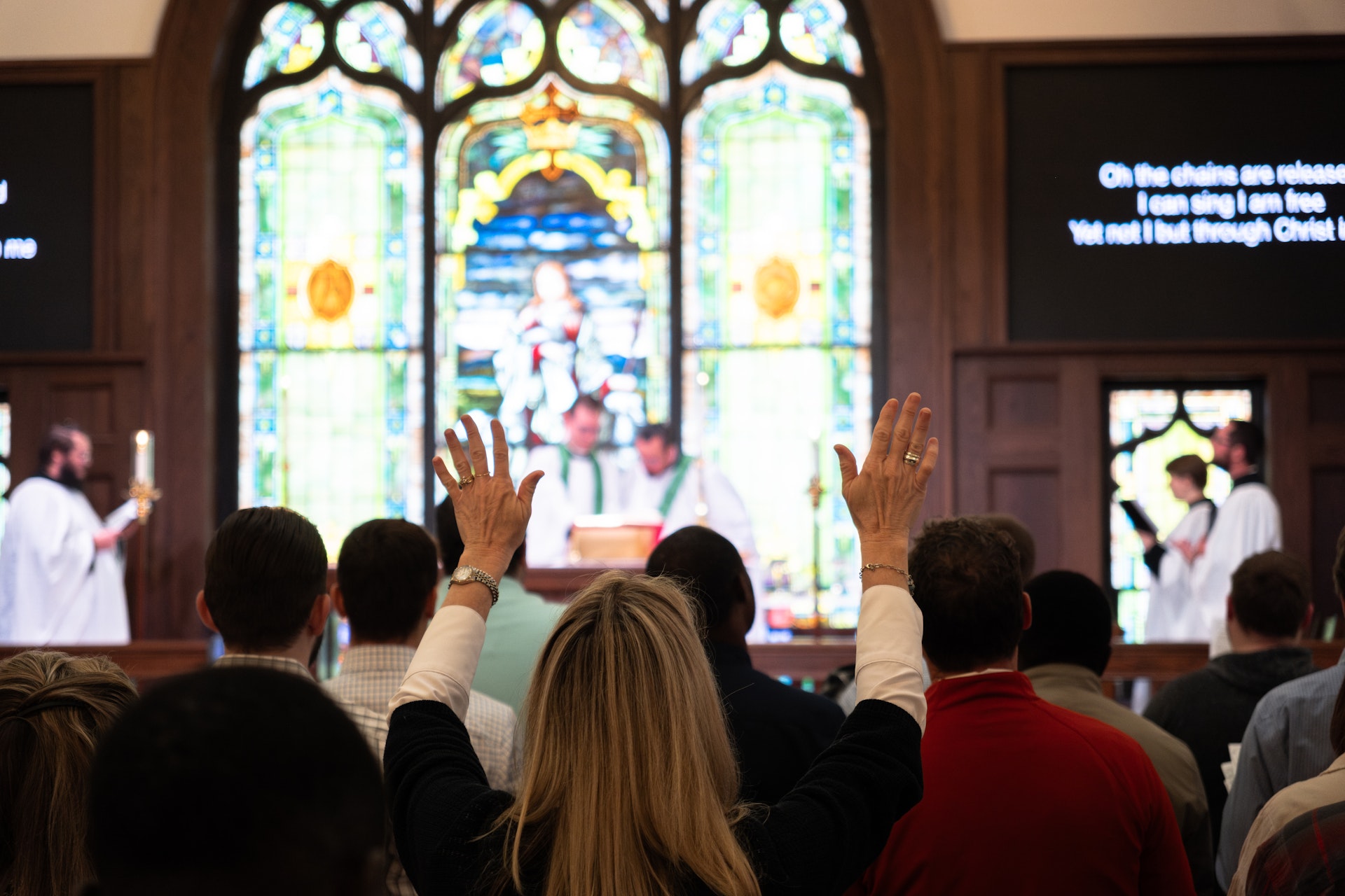 Congregation in church with a woman raising her hands in worship, stained glass windows and clergy at the altar.