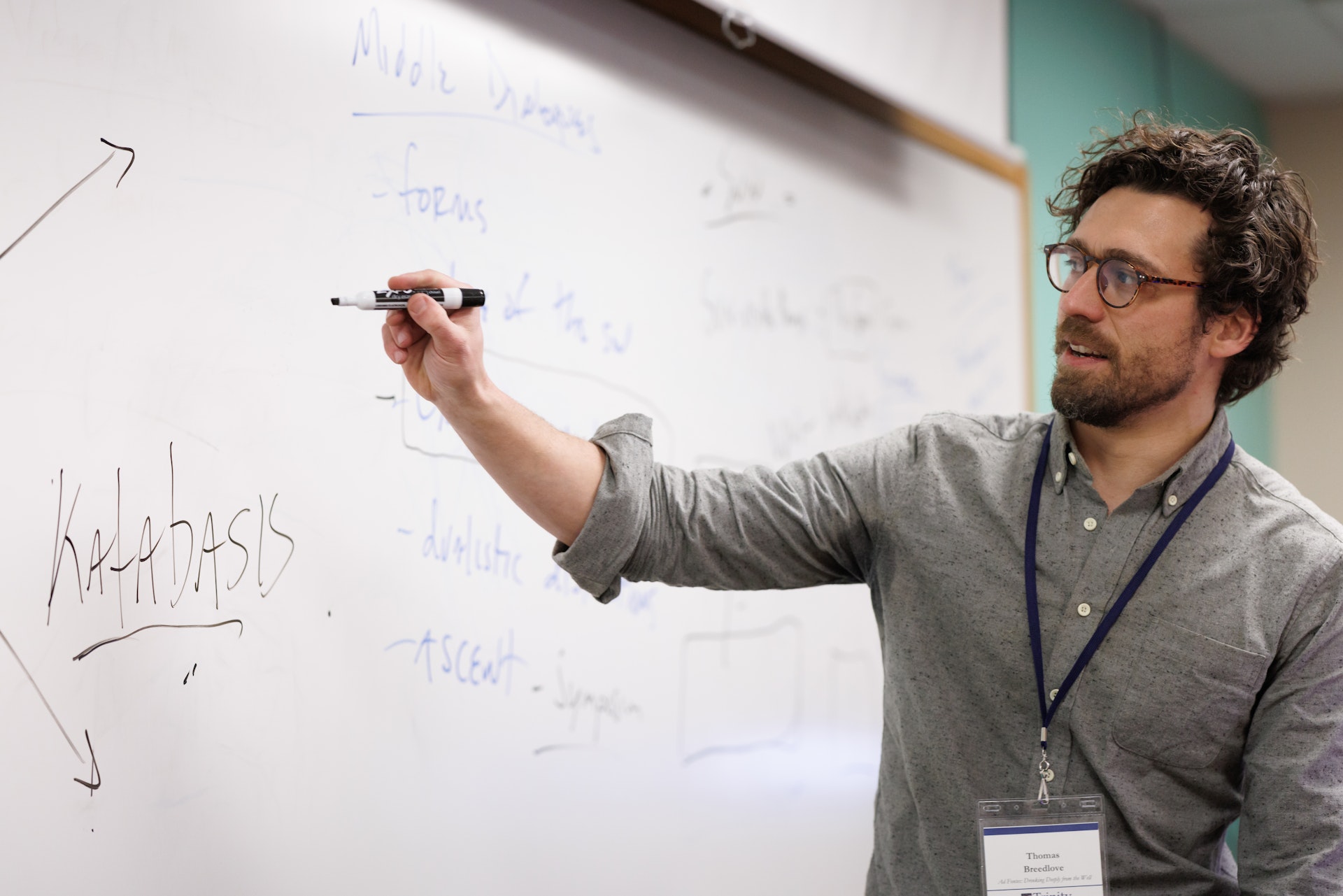 Man with glasses and curly hair writing the word 'Katabasis' on a whiteboard during a lecture.