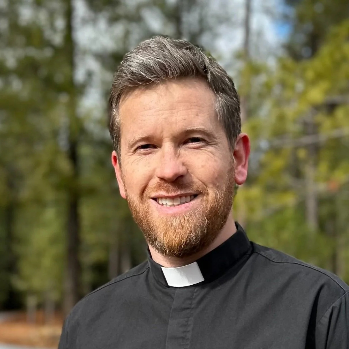 Smiling man wearing a black clerical collar shirt outdoors with trees in the background.