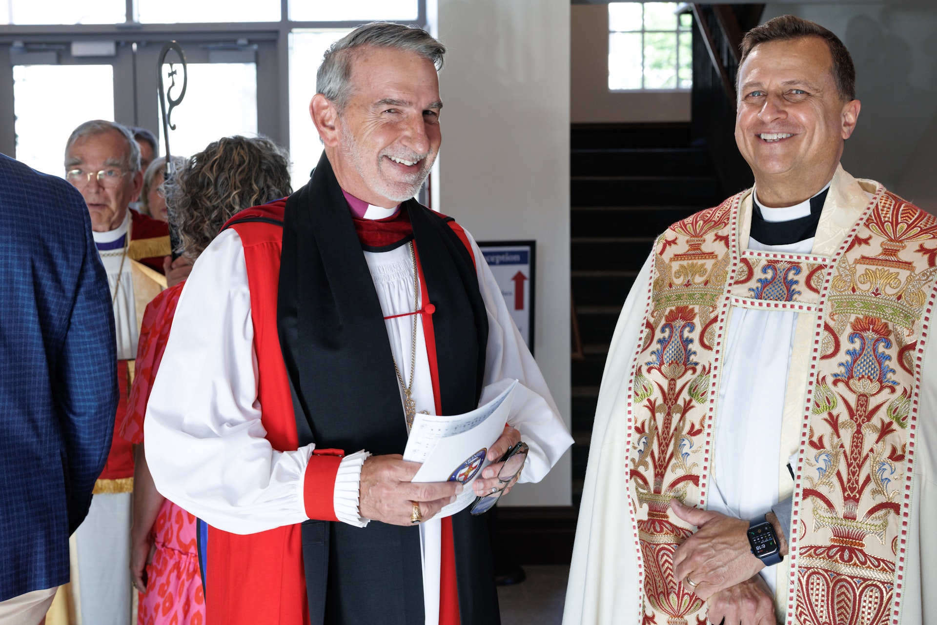Two clergymen smiling indoors, one wearing red and black vestments holding papers and glasses, the other in ornate white and red vestments with a smartwatch.