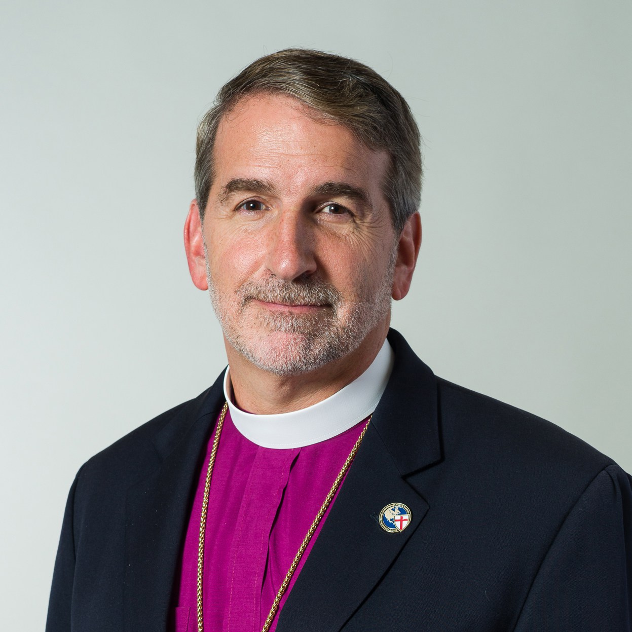 Middle-aged man wearing clerical attire with a purple shirt, white clerical collar, and dark jacket with a religious pin.