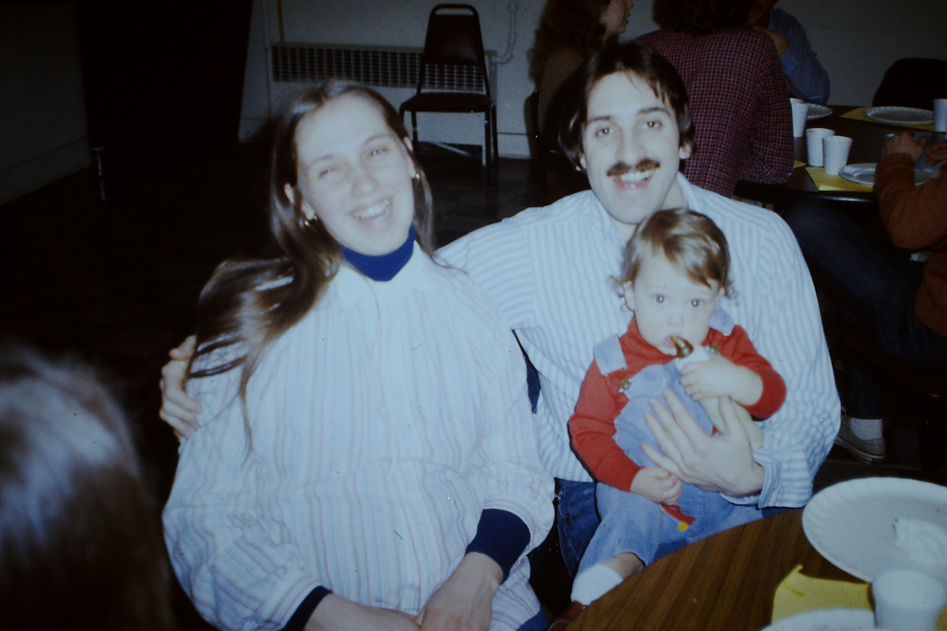 Smiling couple sitting close together, man holding a young child in his lap with people seated and eating at tables in the background.