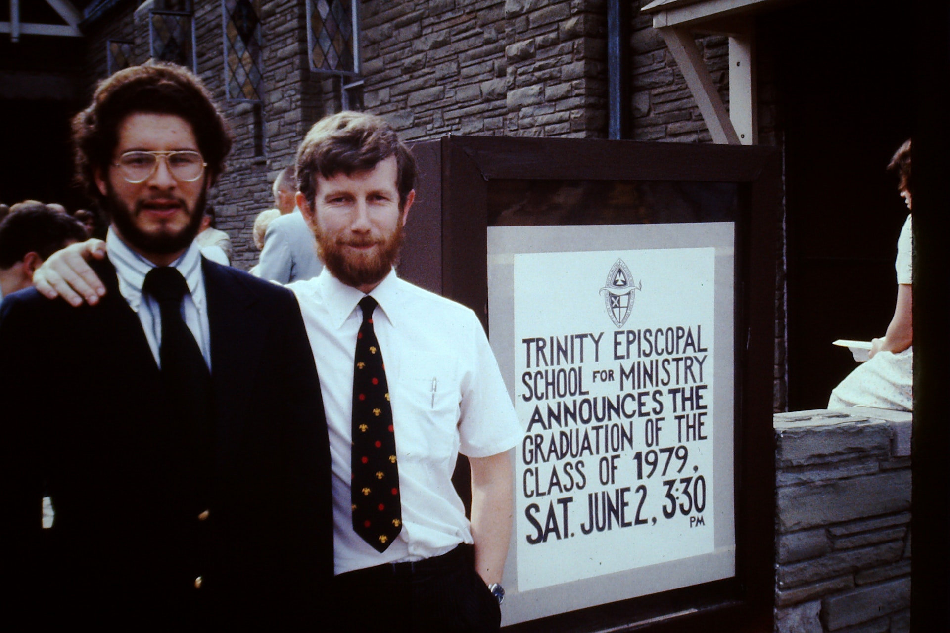 Two men dressed formally standing in front of a sign announcing the 1979 graduation of Trinity Episcopal School for Ministry on June 2 at 3:30 PM.