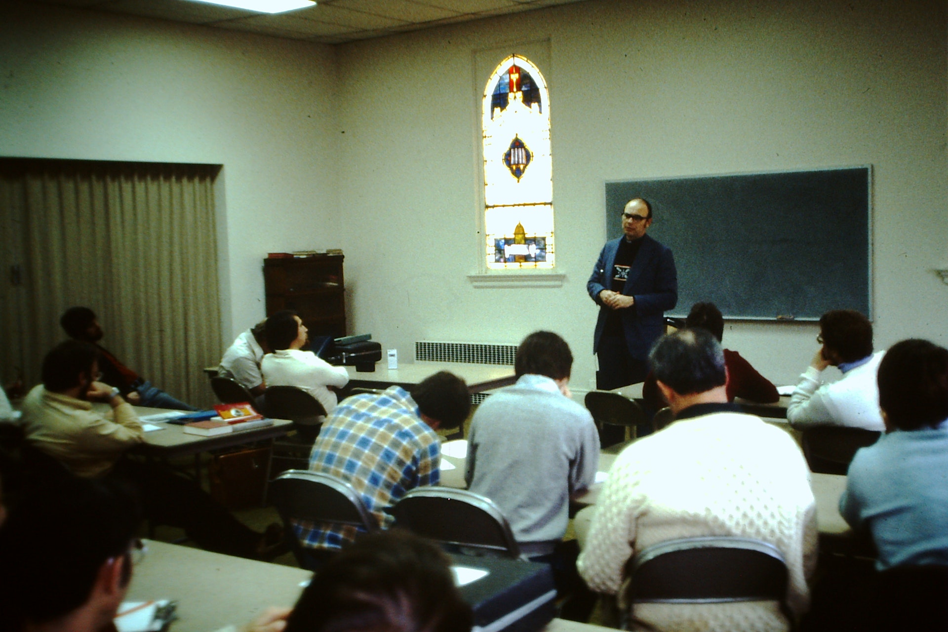 Man standing and speaking in front of a classroom with seated students, a stained glass window, and a chalkboard in the background.