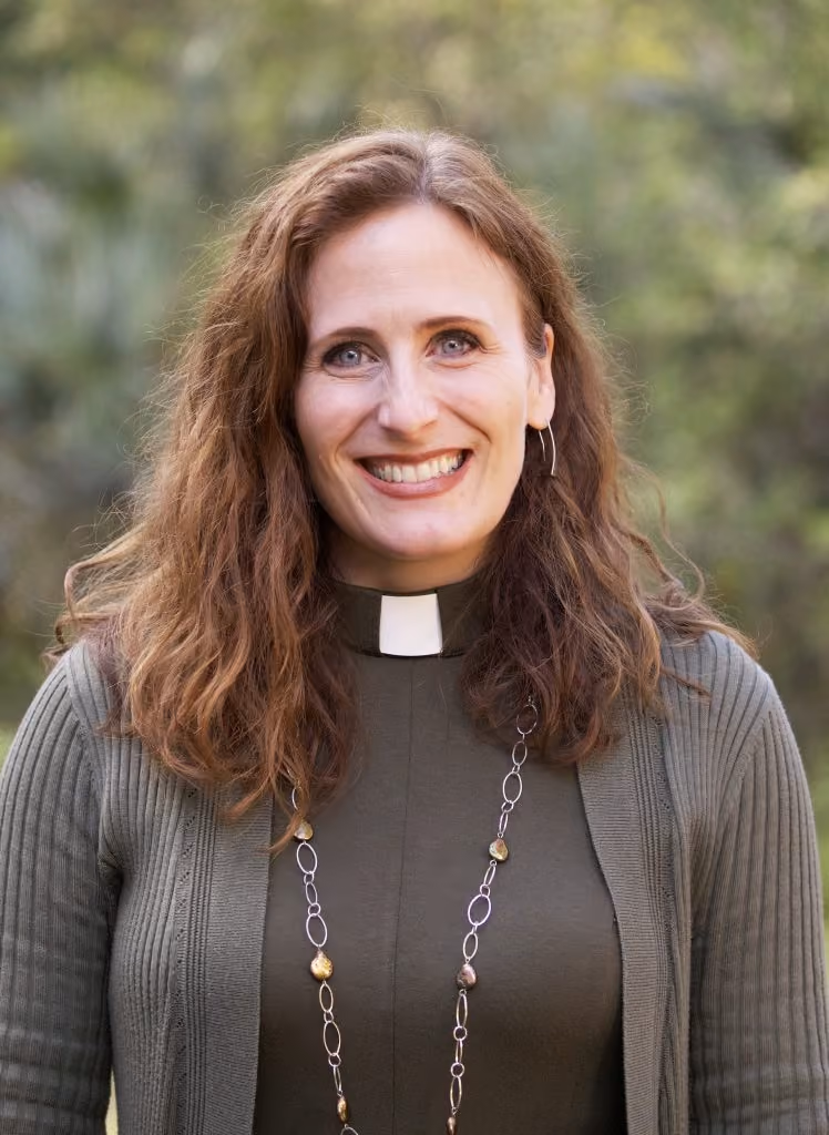 Smiling woman with curly hair wearing clerical collar, necklace, and green cardigan outdoors.